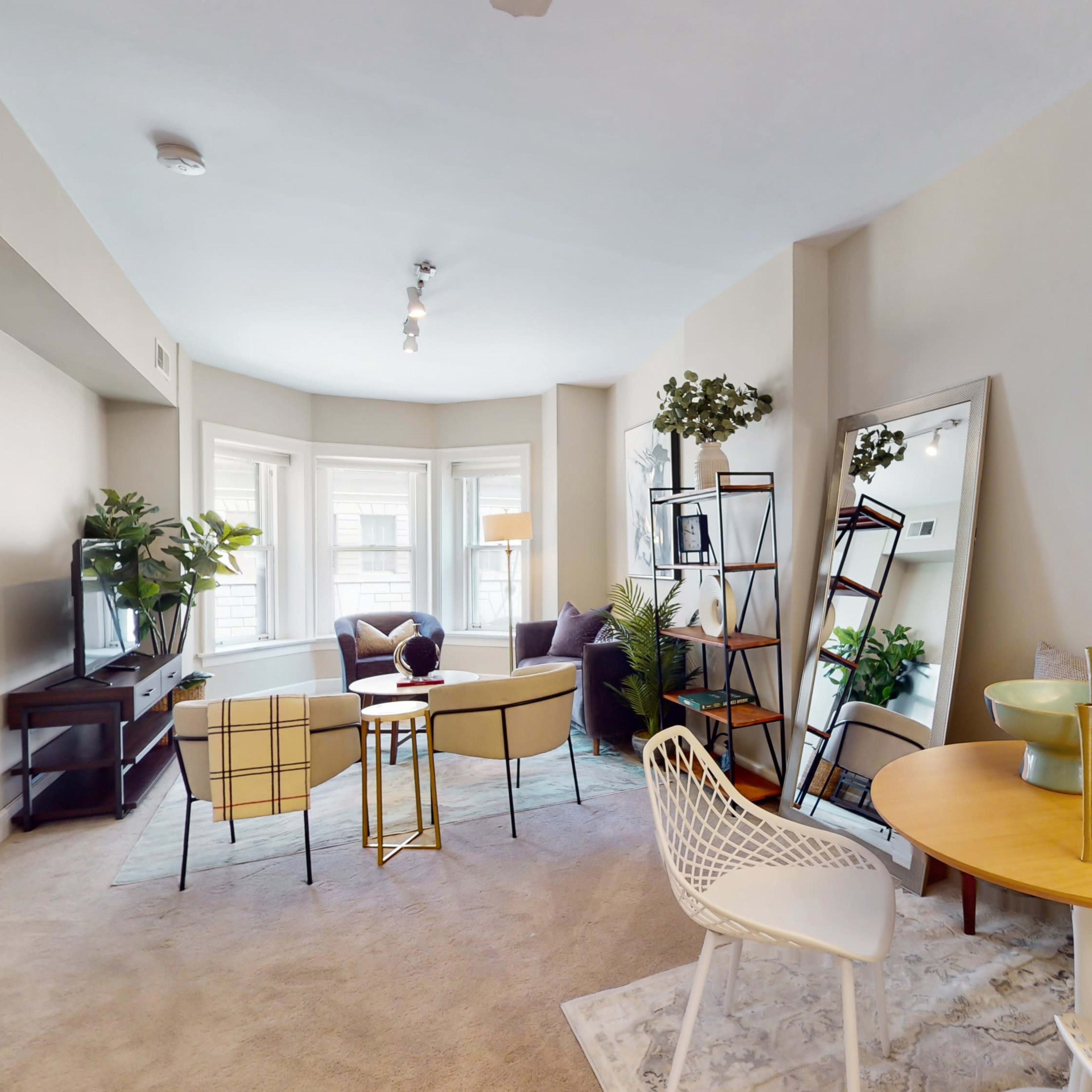 A well-furnished living space featuring a seating area with a round table, a television stand, and large windows with natural light, surrounded by decorative plants and a mirror.