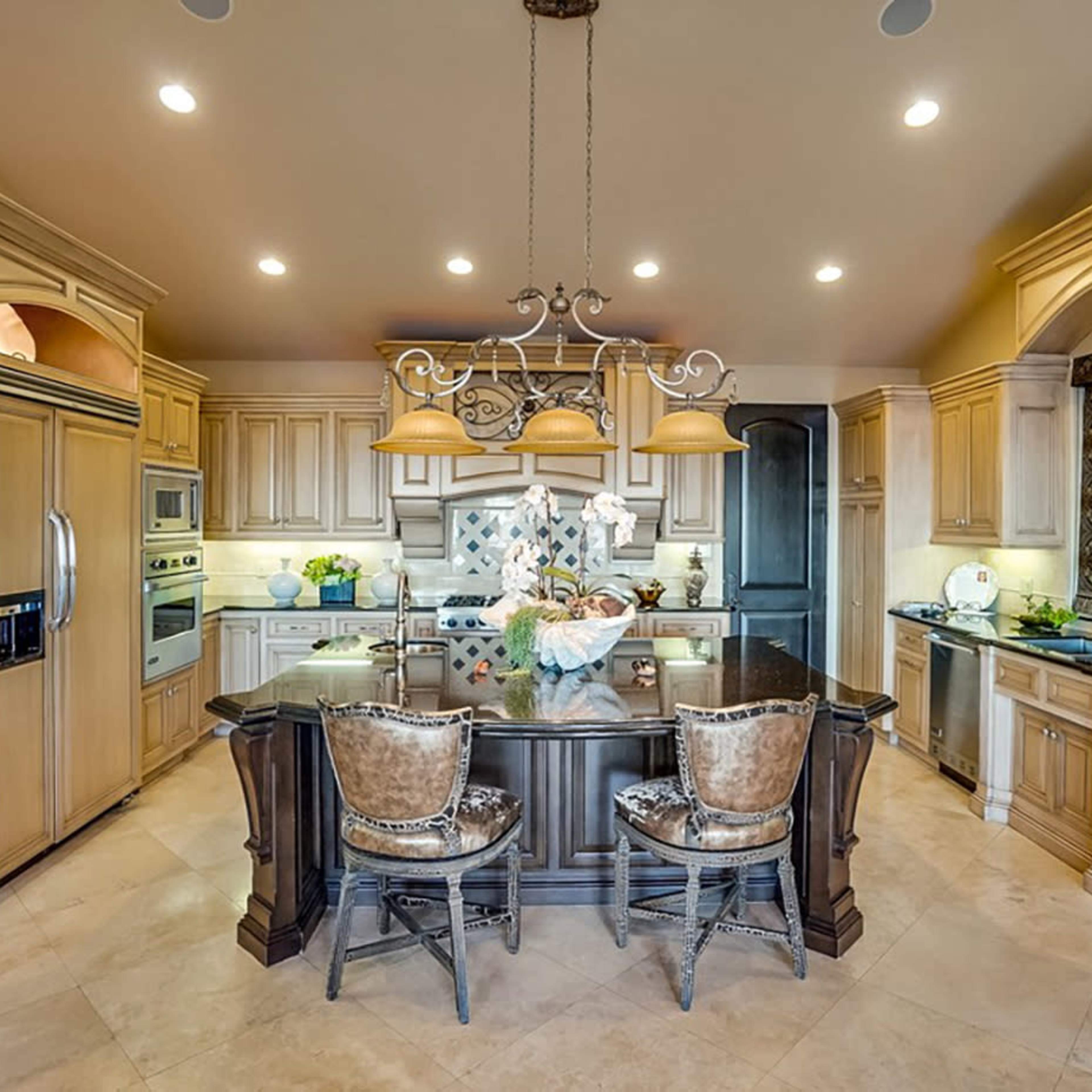 A spacious kitchen features light wood cabinetry, a large island with dark counters, and elegant bar stools, along with modern appliances and decorative accents.
