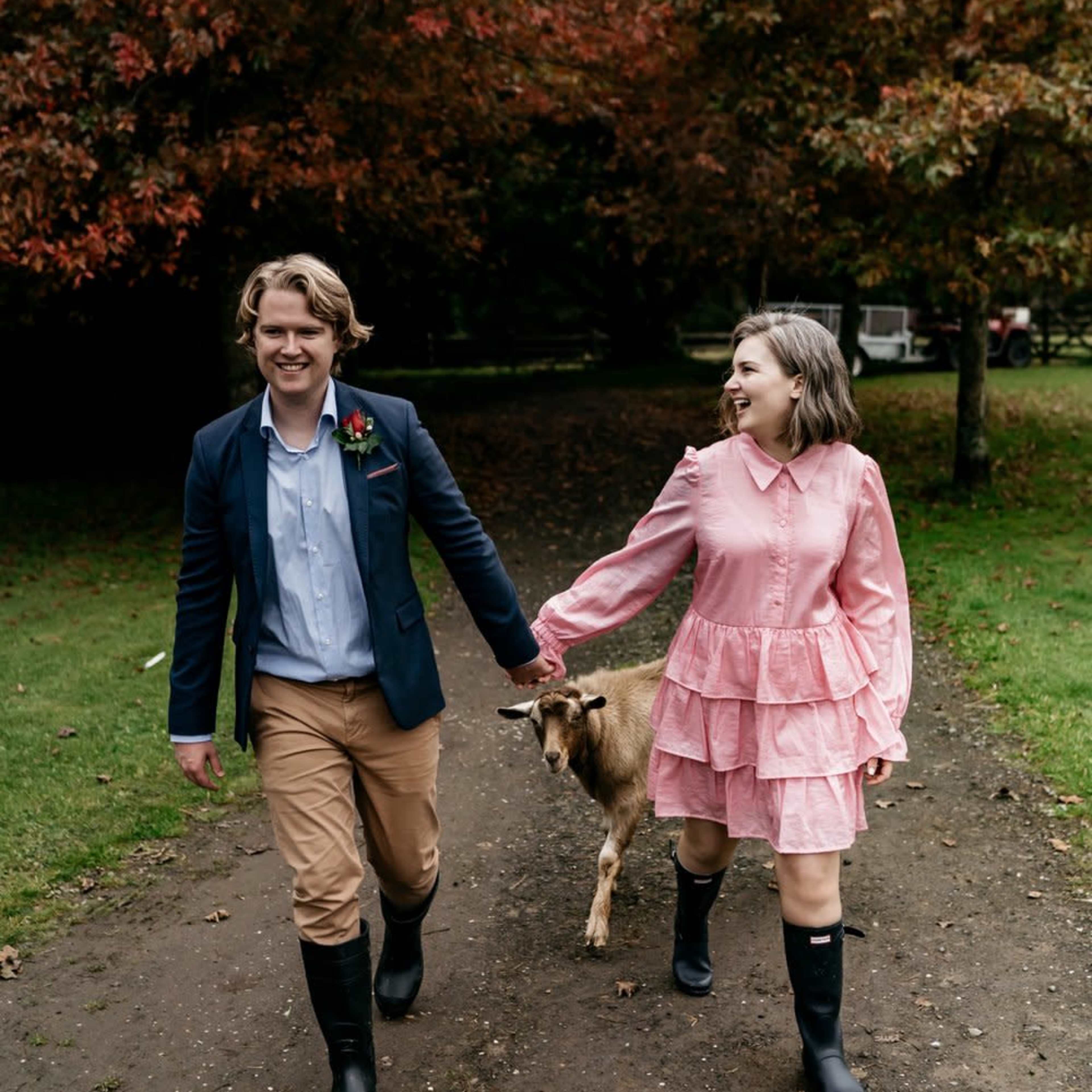 A couple walks hand-in-hand with a goat along a dirt path surrounded by trees with autumn foliage.