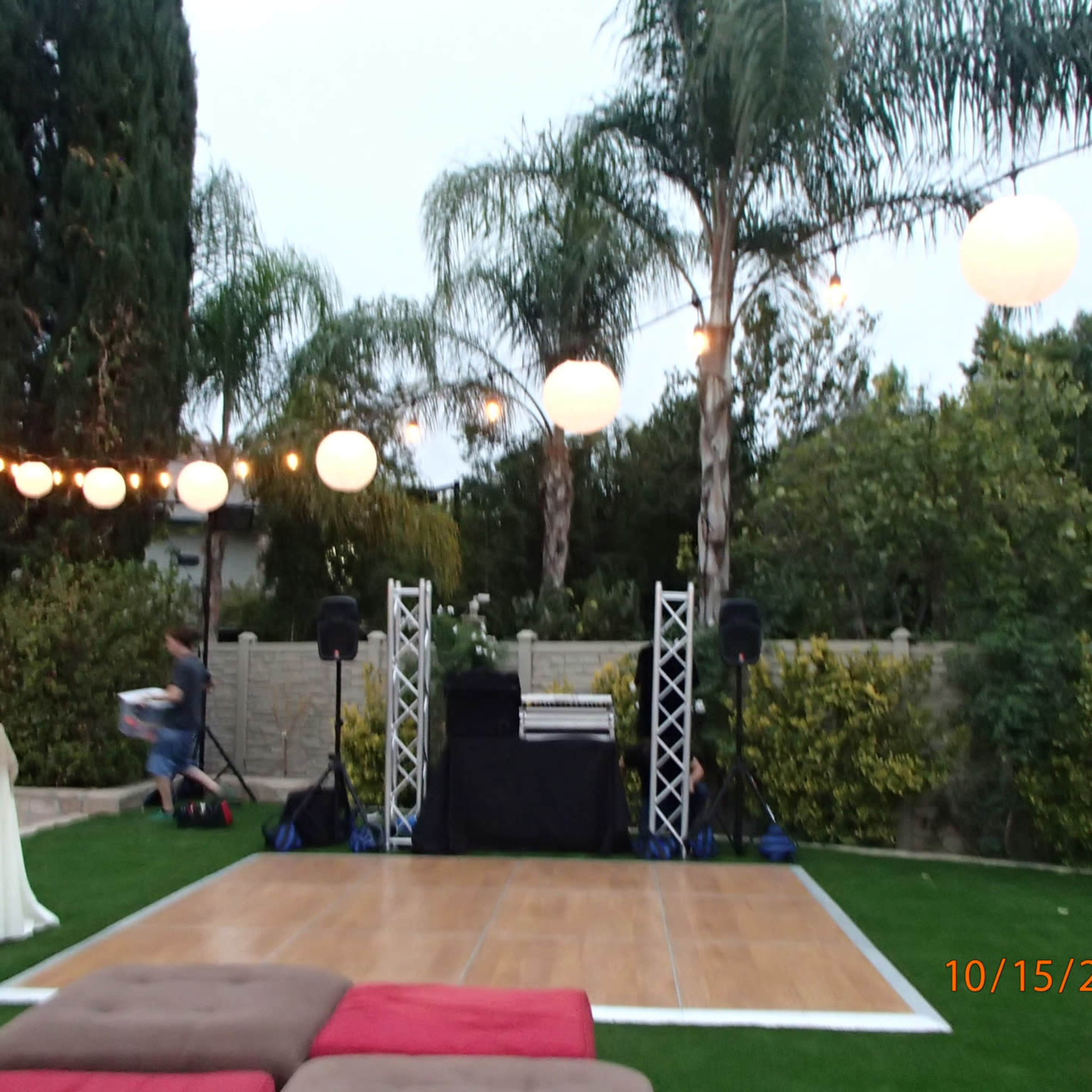 An outdoor event space with a wooden dance floor, illuminated by hanging lanterns and surrounded by palm trees.