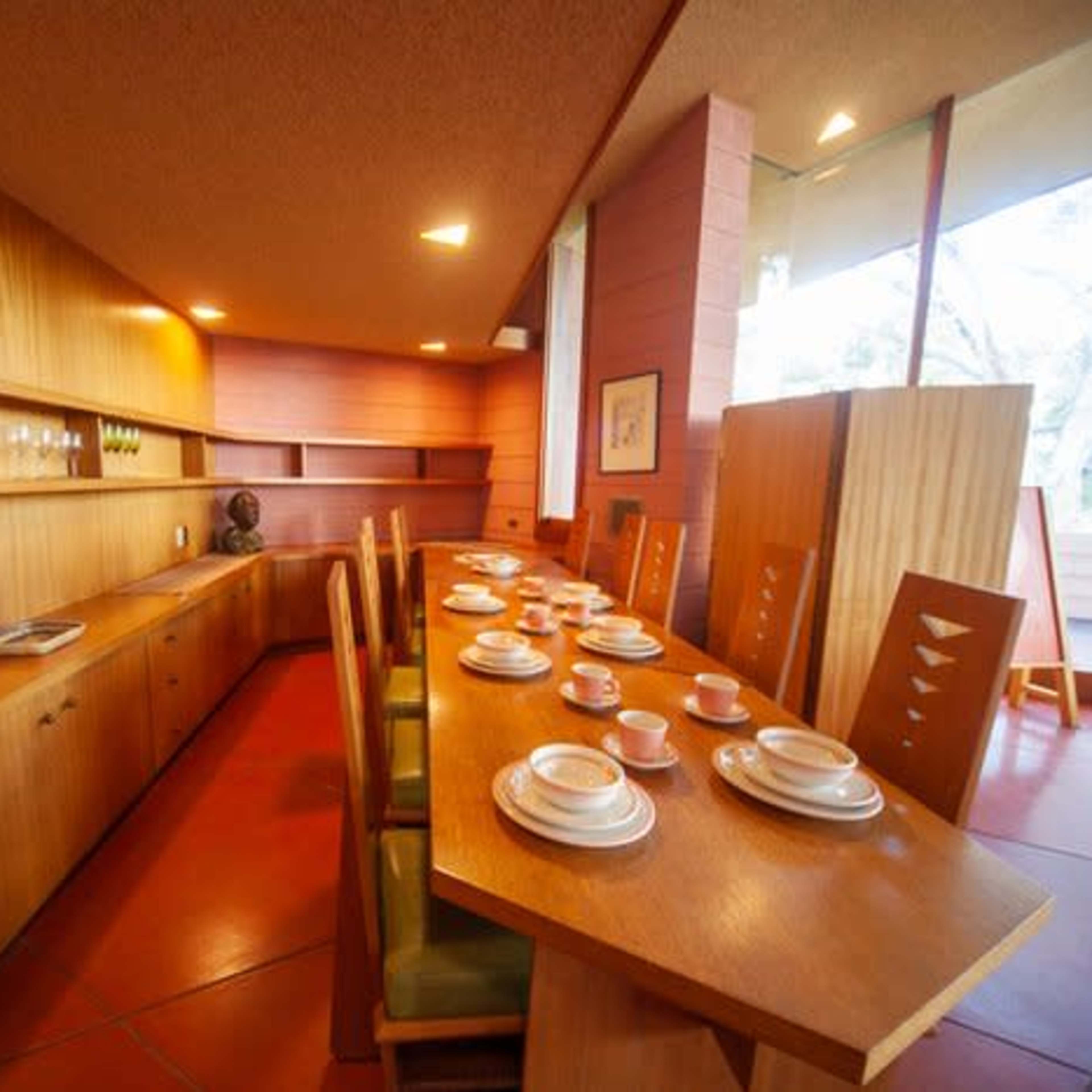 A modern dining area features a wooden table set with plates and cups, surrounded by green chairs against a backdrop of wooden cabinetry and large windows.