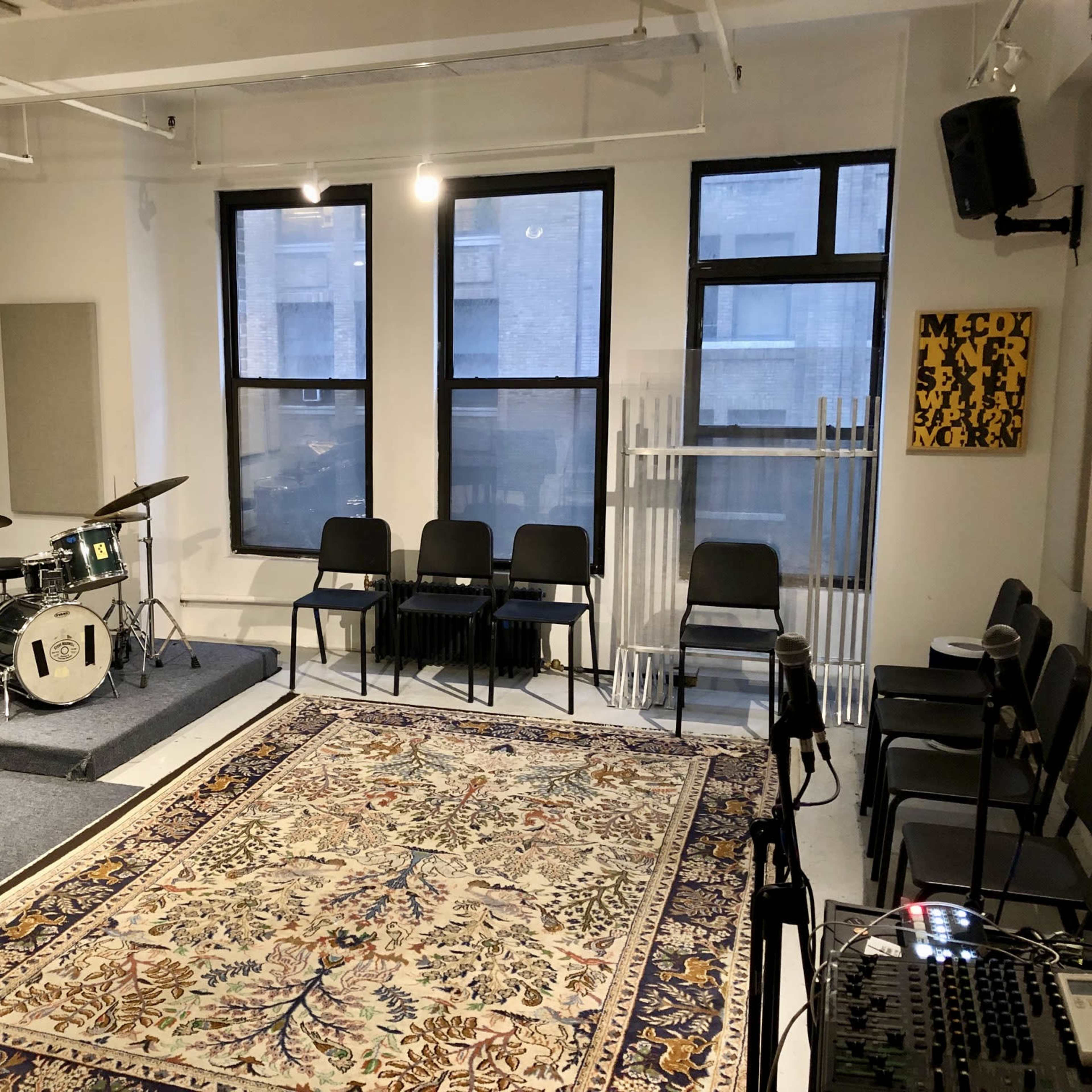 A music rehearsal studio featuring a drum set, several chairs arranged in a row, and a large patterned rug on the floor.