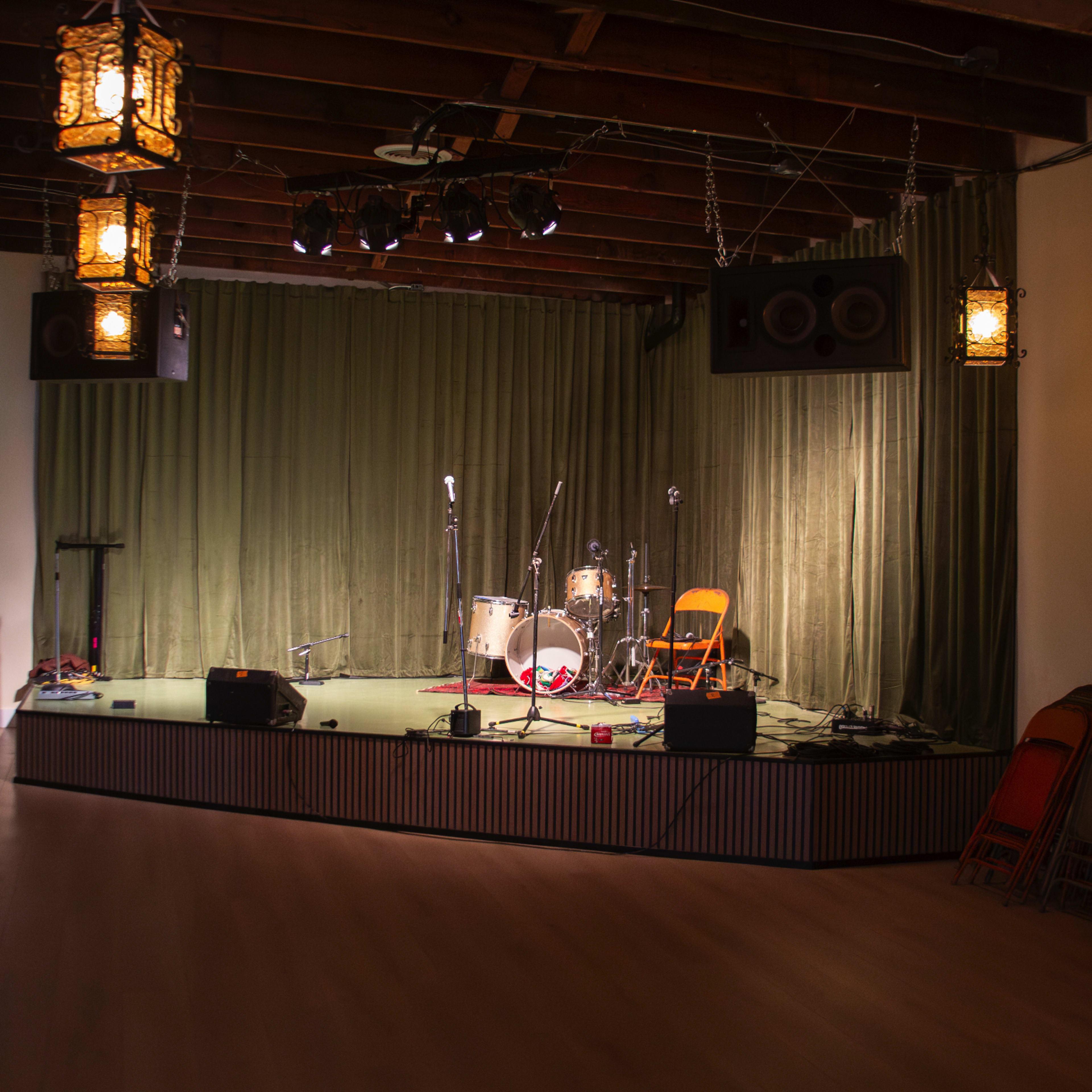 A small stage is set up with musical equipment and a single orange chair, surrounded by green curtains and overhead lanterns.