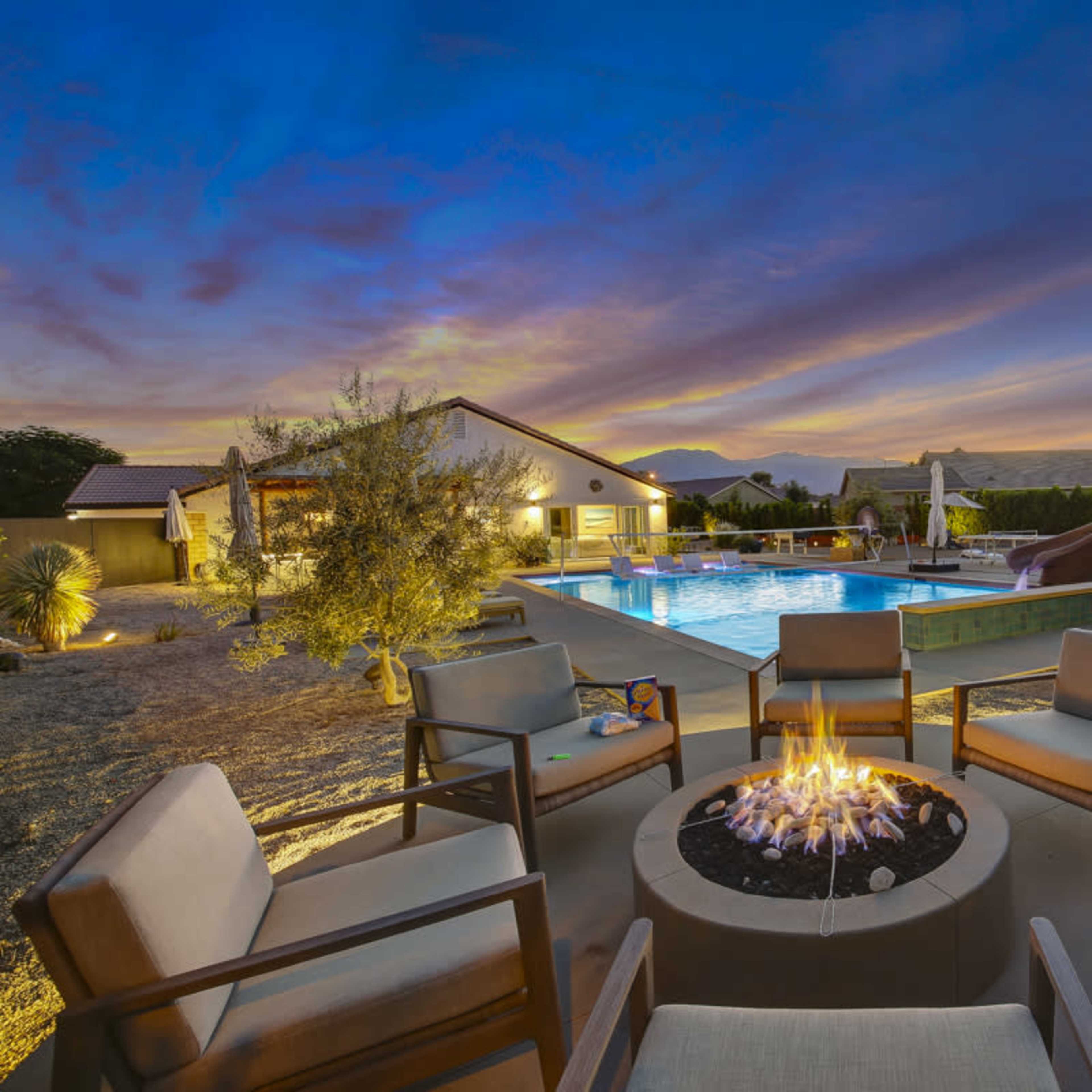 A circular fire pit with chairs surrounds a pool area that features a water slide and illuminated landscaping against a twilight sky with mountains in the background.