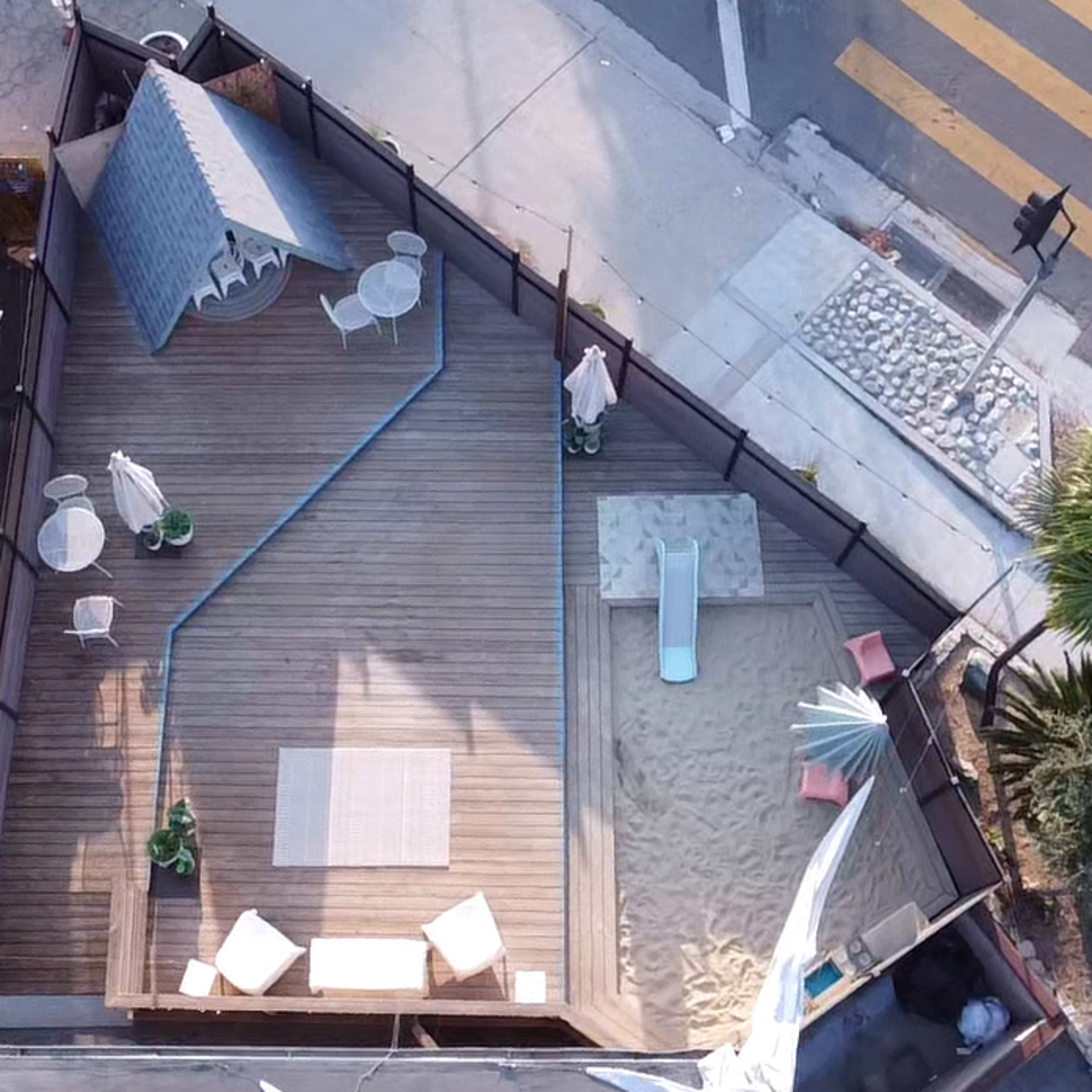 An aerial view of a wooden deck area with lounge furniture, a small pool, and a sand play area, all enclosed by a fence near a street intersection.