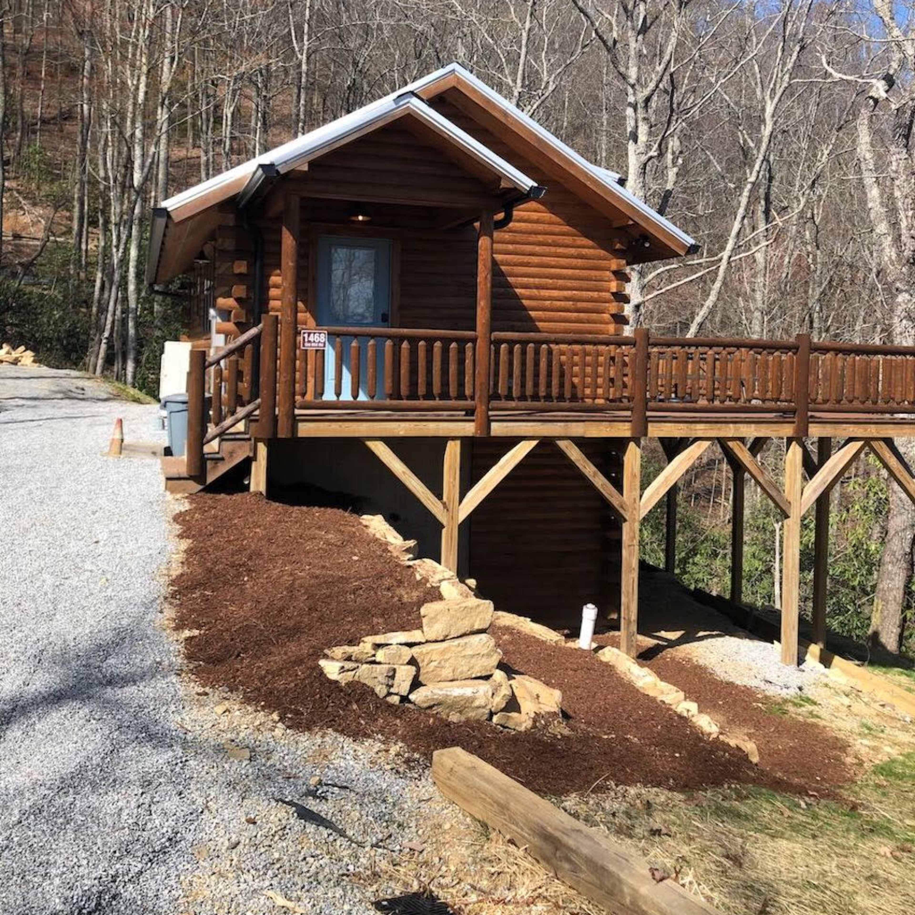 A wooden cabin with a wraparound porch is situated on a gravel driveway surrounded by trees.