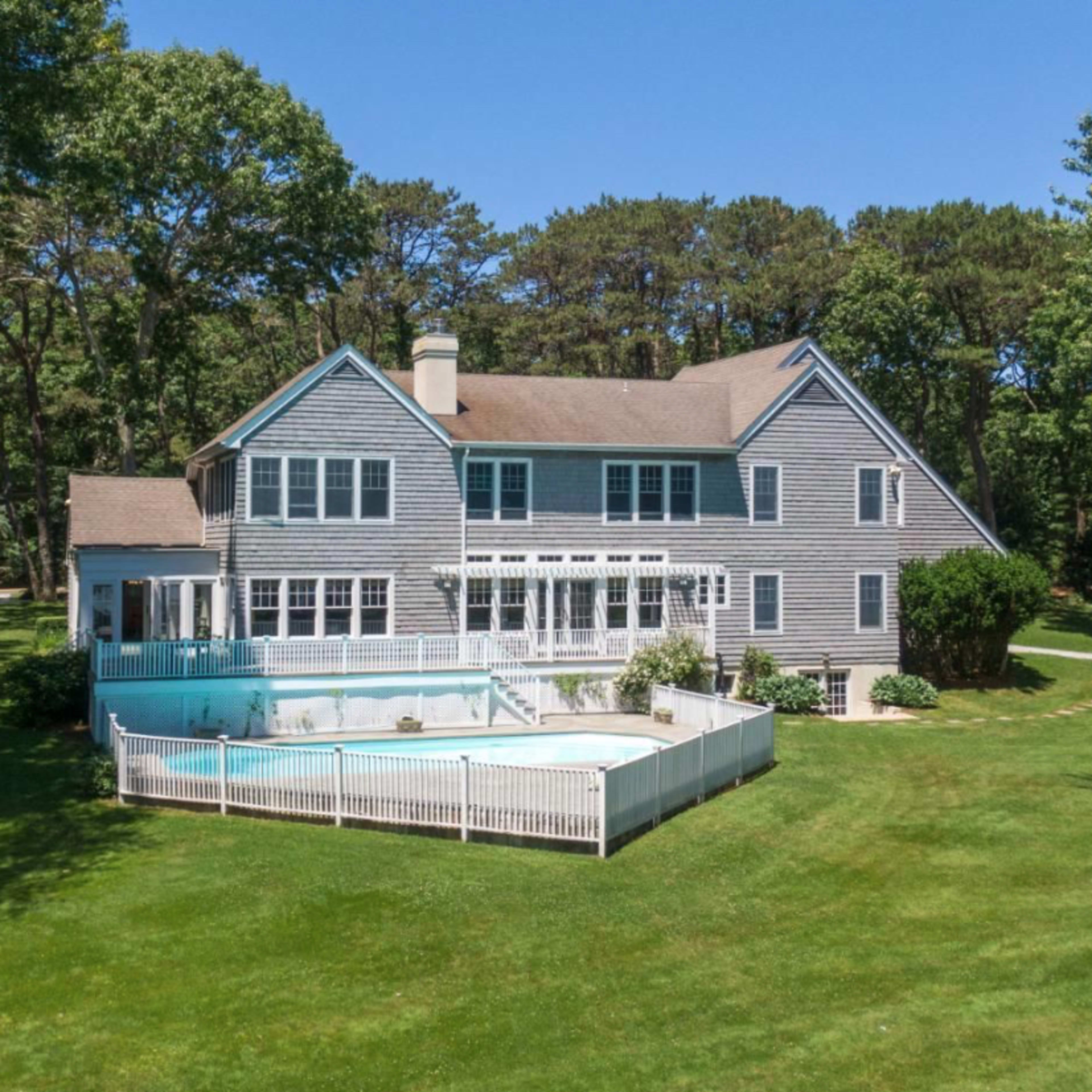 A large gray house with multiple windows and a swimming pool surrounded by a white fence sits on a grassy lawn, bordered by trees.