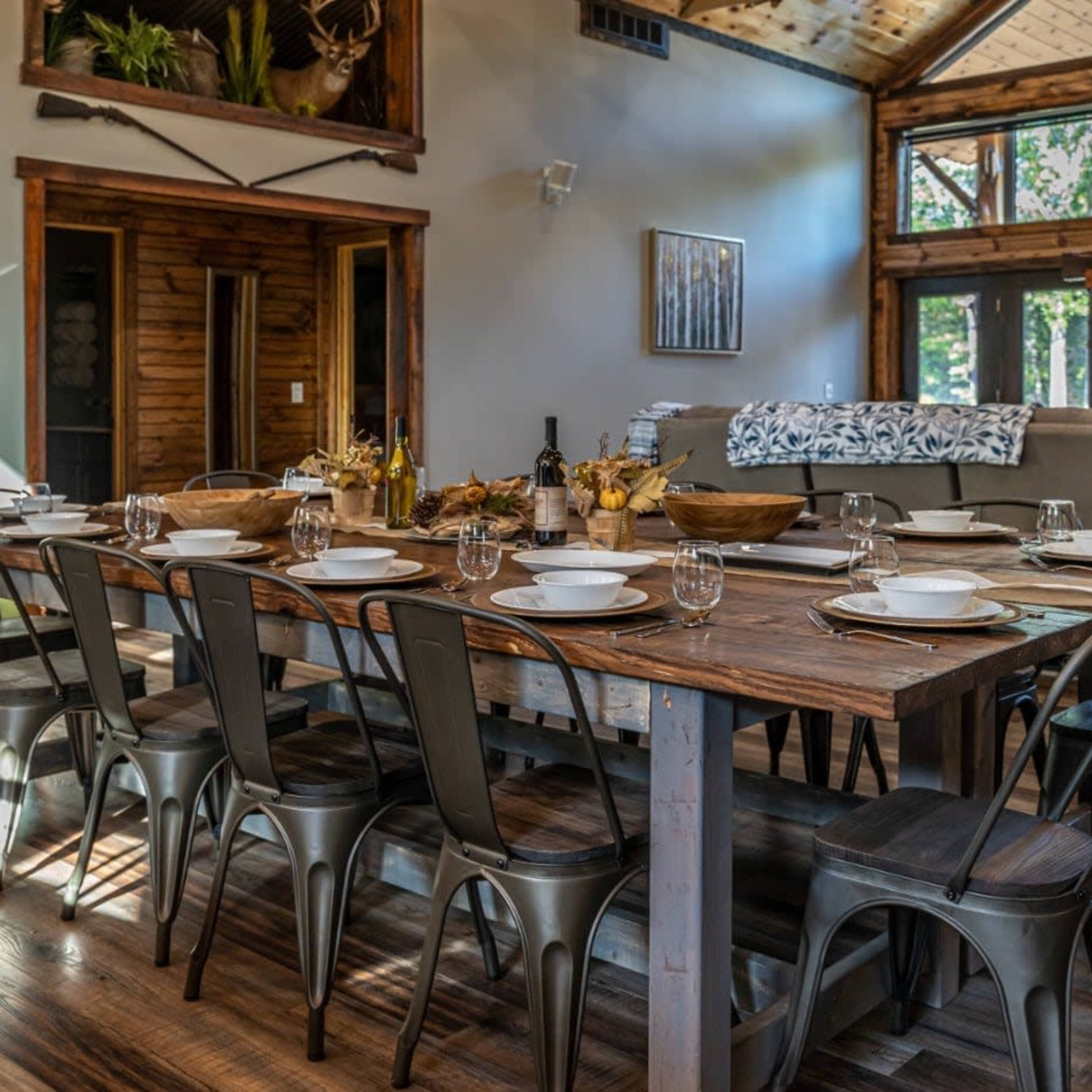 A long wooden dining table is set with plates and glassware in a rustic, well-lit room featuring large windows and a stone fireplace.