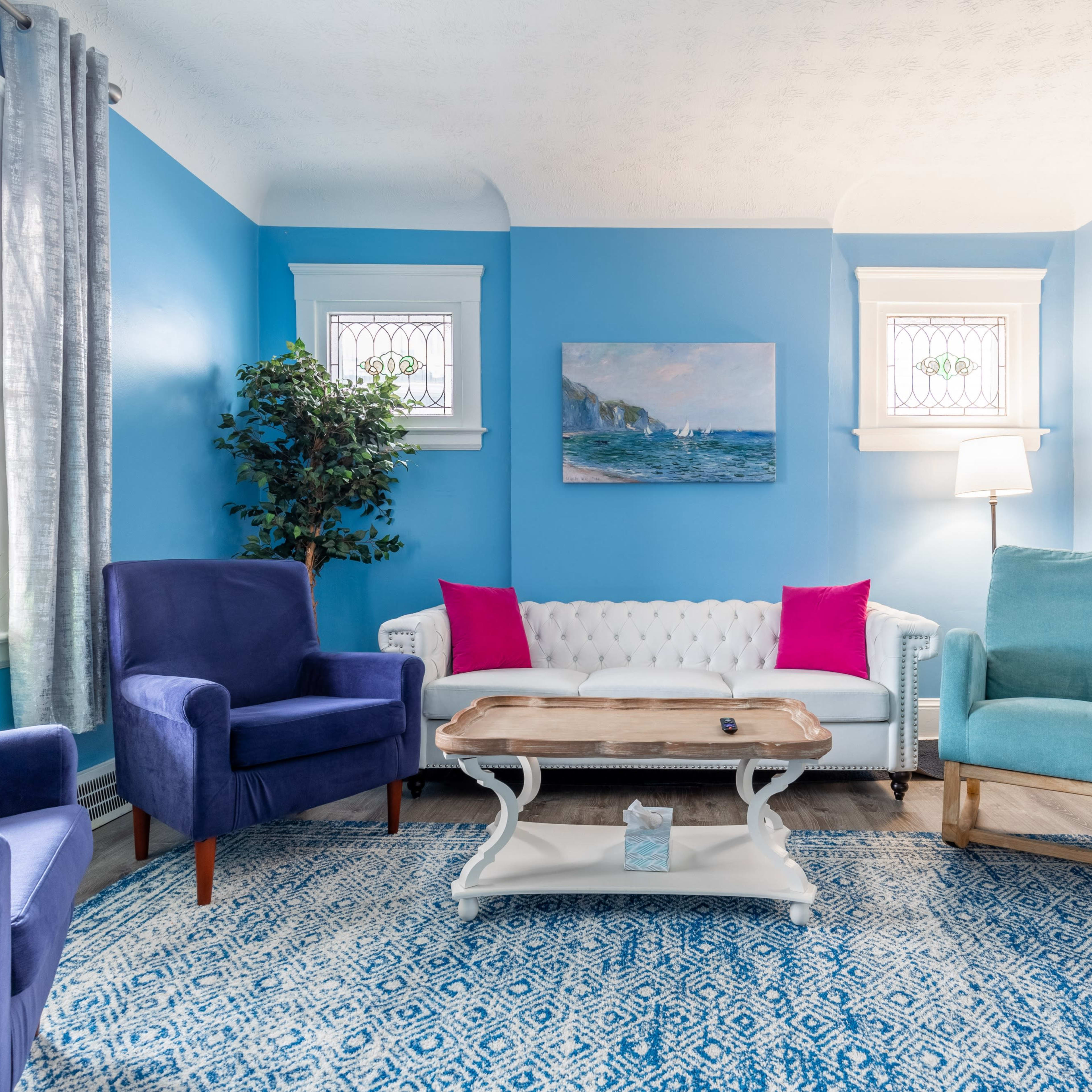 A brightly colored living room with blue walls, a white couch, and various seating options arranged around a wooden coffee table on a patterned rug.