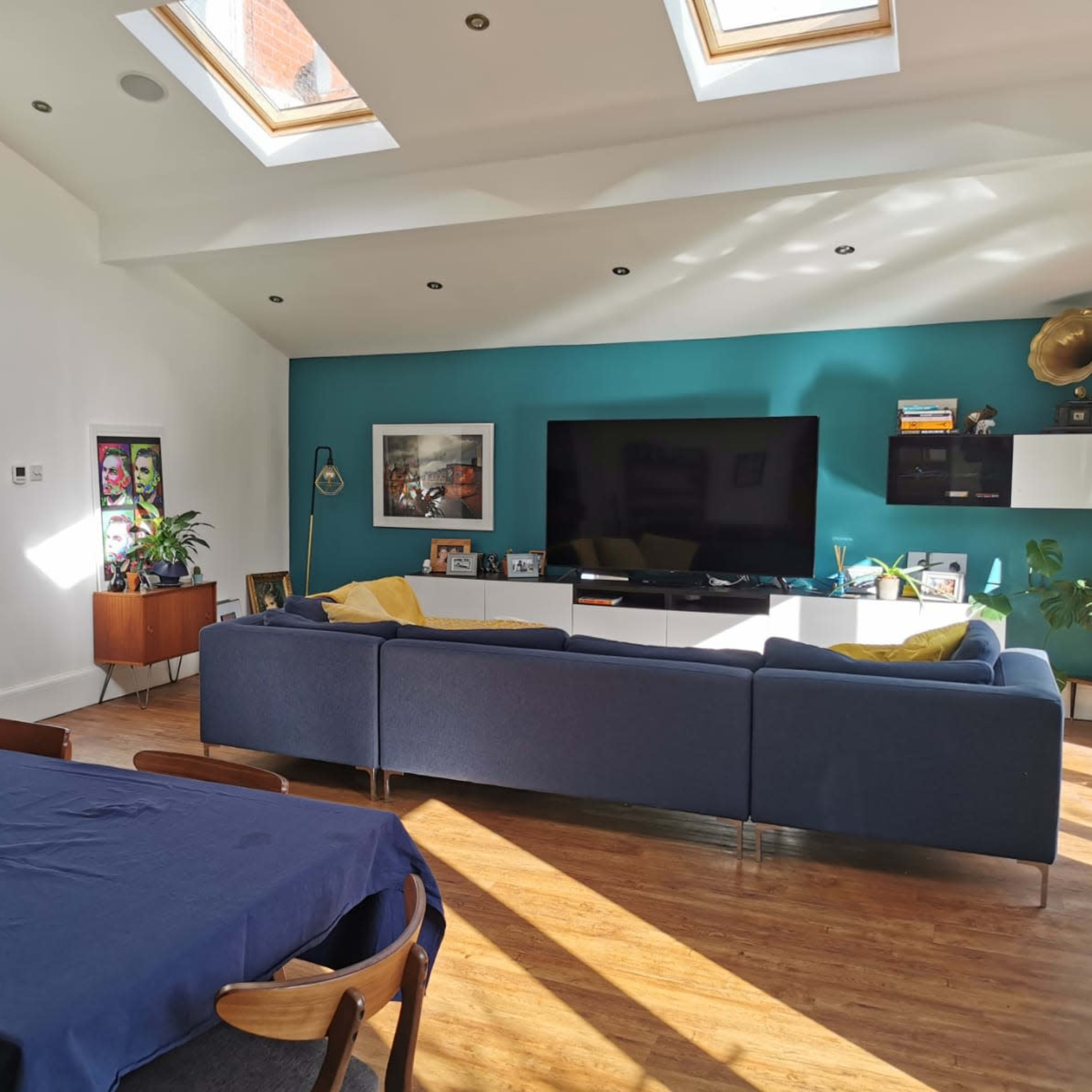 A modern living space with a blue sofa, a large television against a teal wall, a dining table, and skylights overhead.