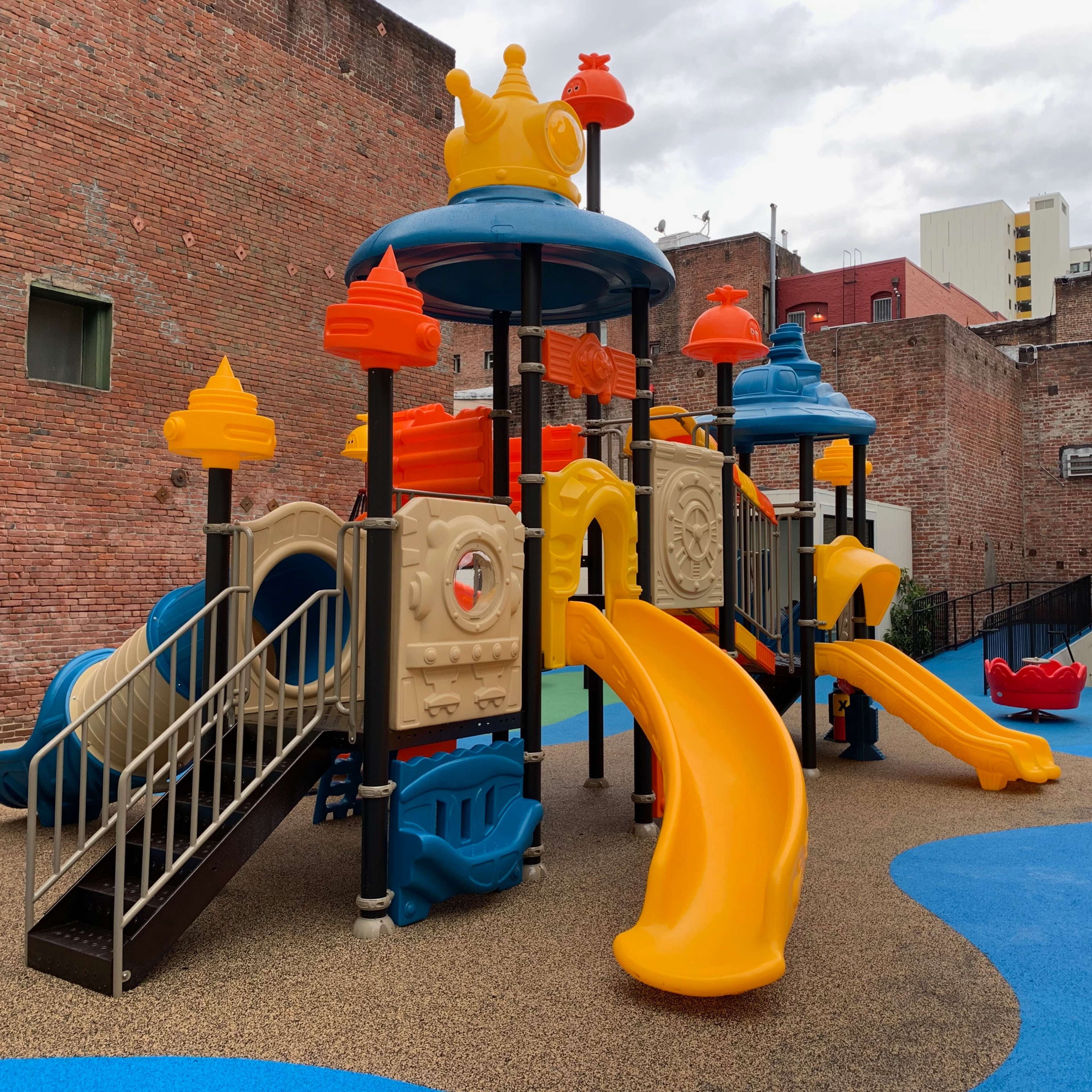 A brightly colored playground structure with slides and climbing features situated on a blue surface in an urban setting.