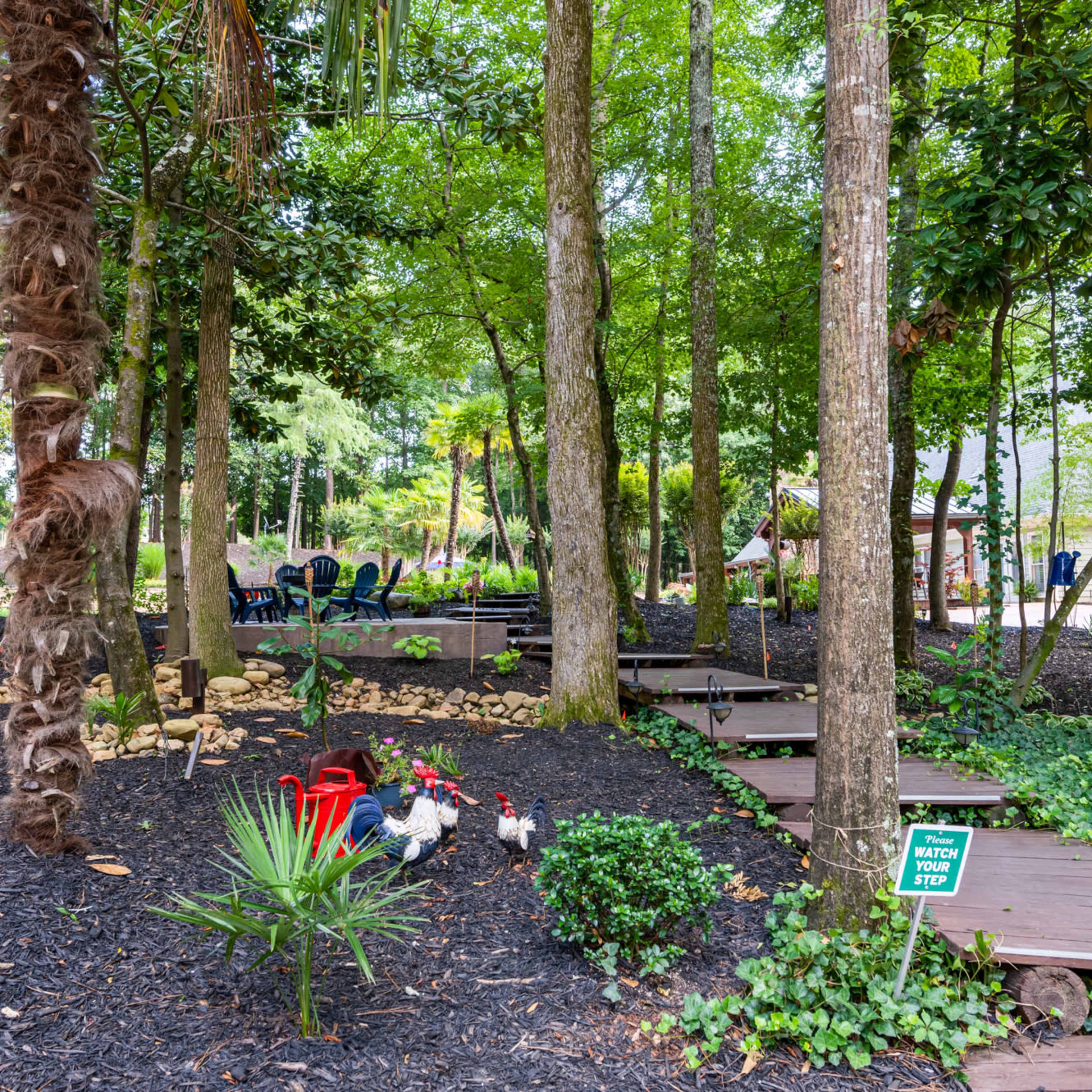 The scene features a landscaped backyard with wooden pathways, surrounded by trees and decorative plants, including seating areas and garden ornaments.