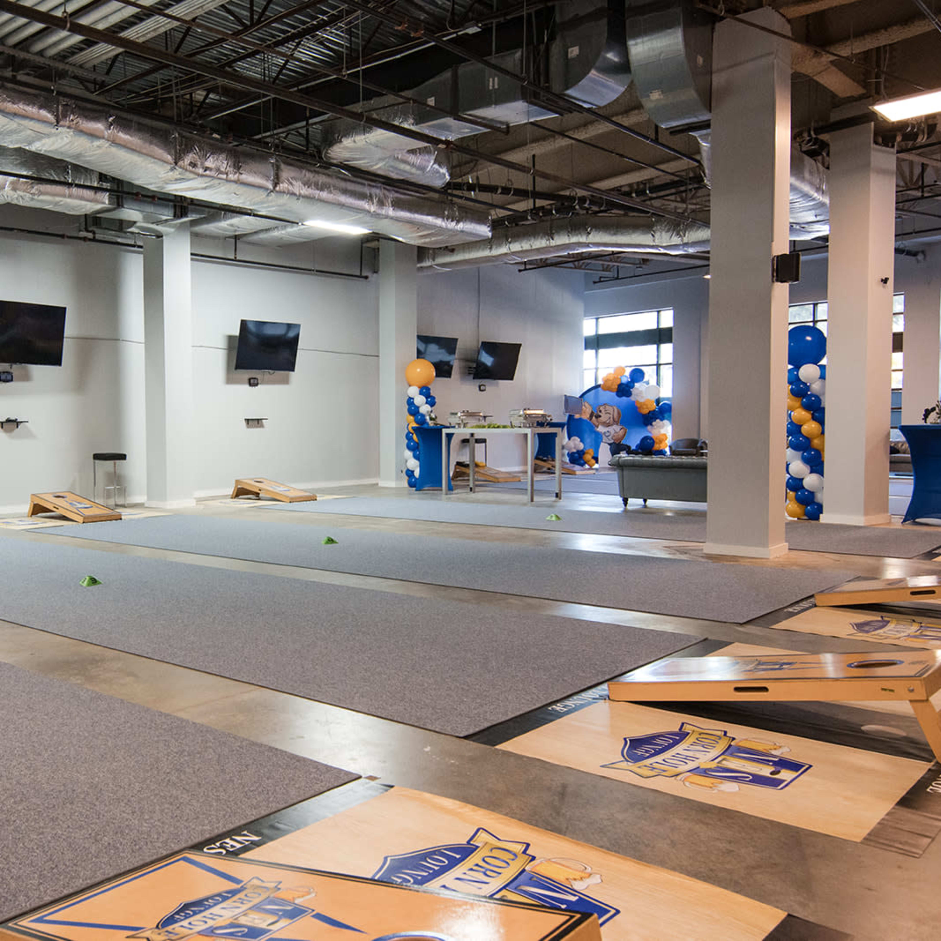 A spacious indoor area set up for cornhole games, featuring several cornhole boards on a carpeted floor and multiple screens mounted on the walls.