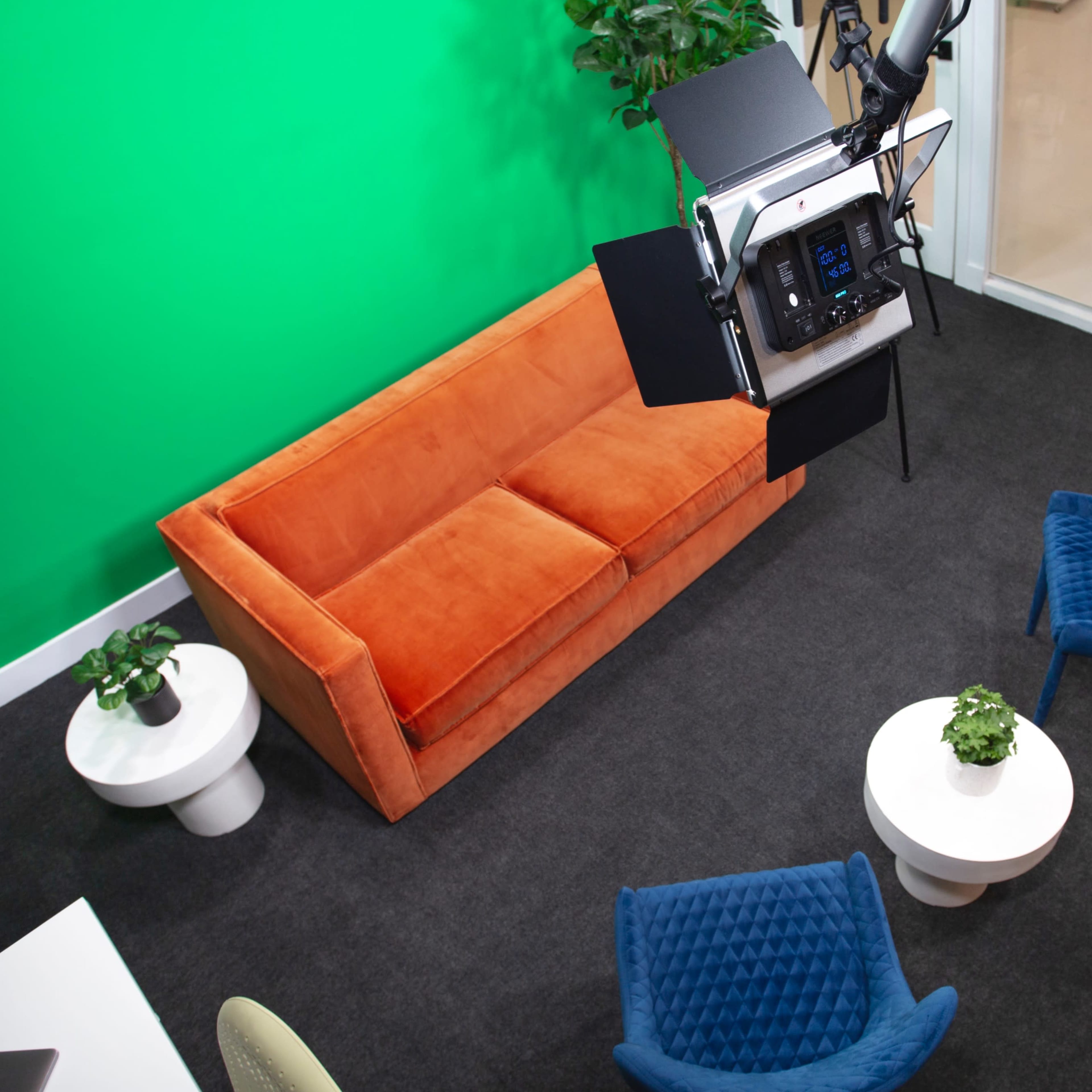A modern studio setup with a green screen, an orange sofa, blue chairs, and a lighting rig overhead.