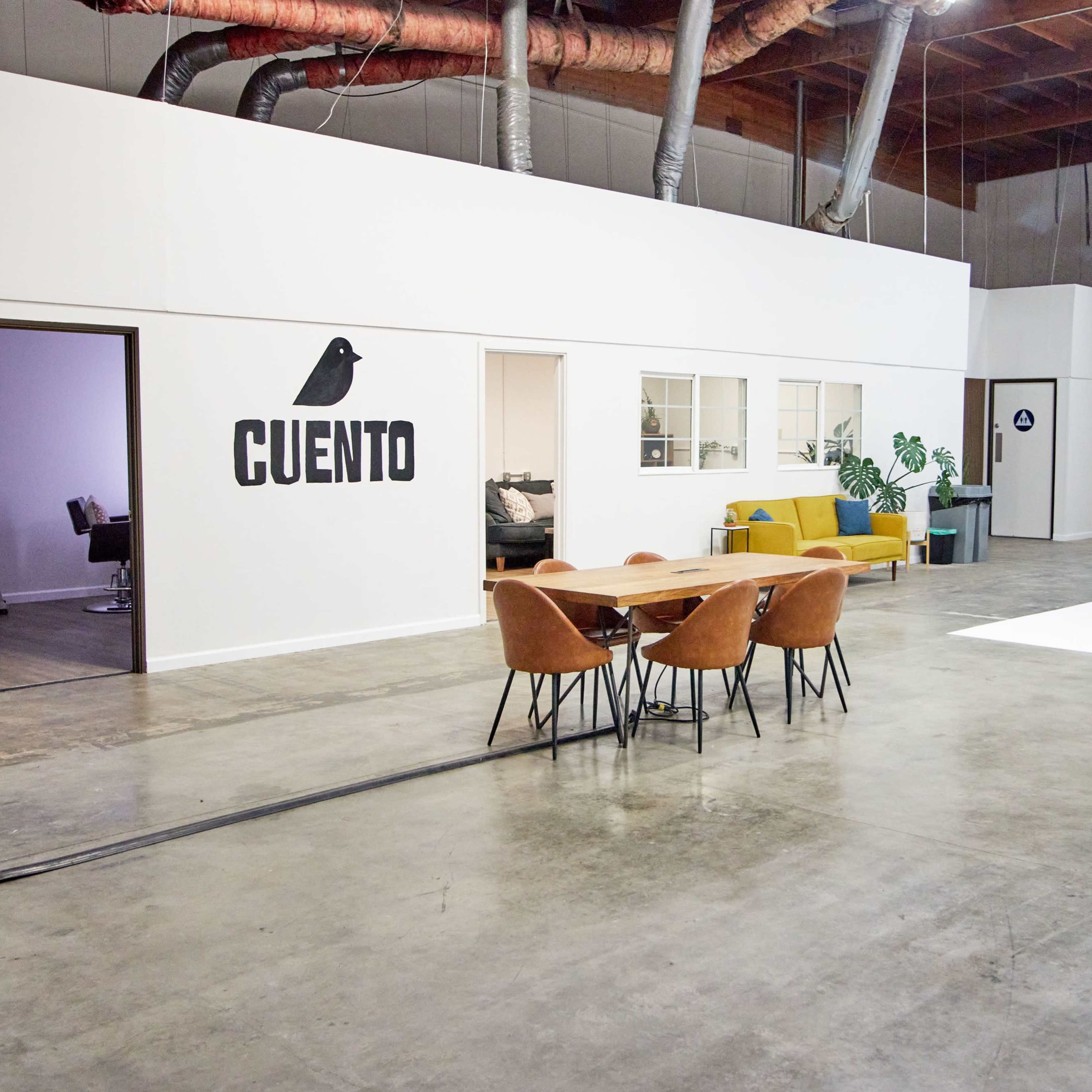 A modern studio space featuring a dining table with chairs, a sofa, and a white backdrop area, with the word "CUENTO" displayed on the wall.