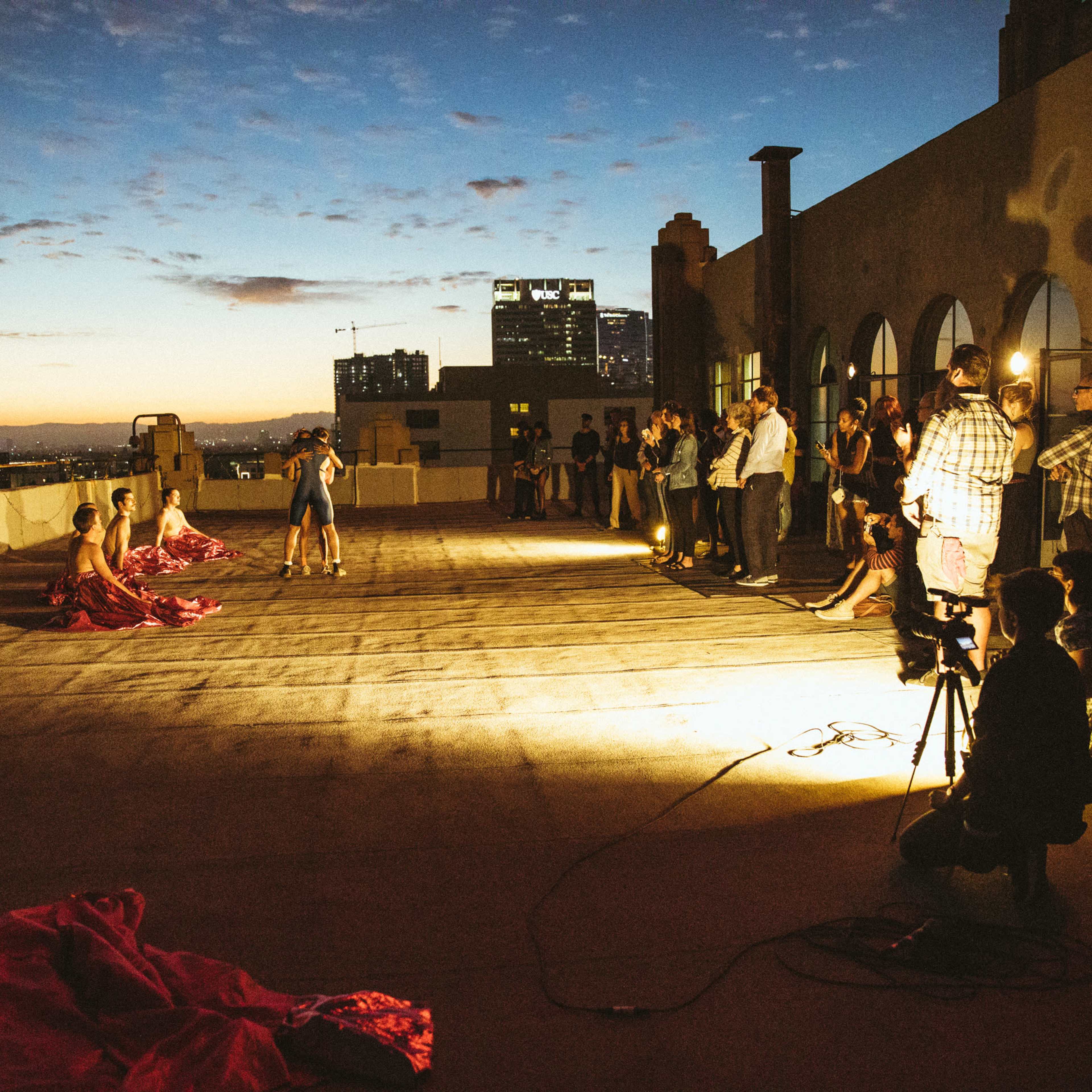 The scene depicts a rooftop performance with dancers in pink fabric as an audience watches under a twilight sky.