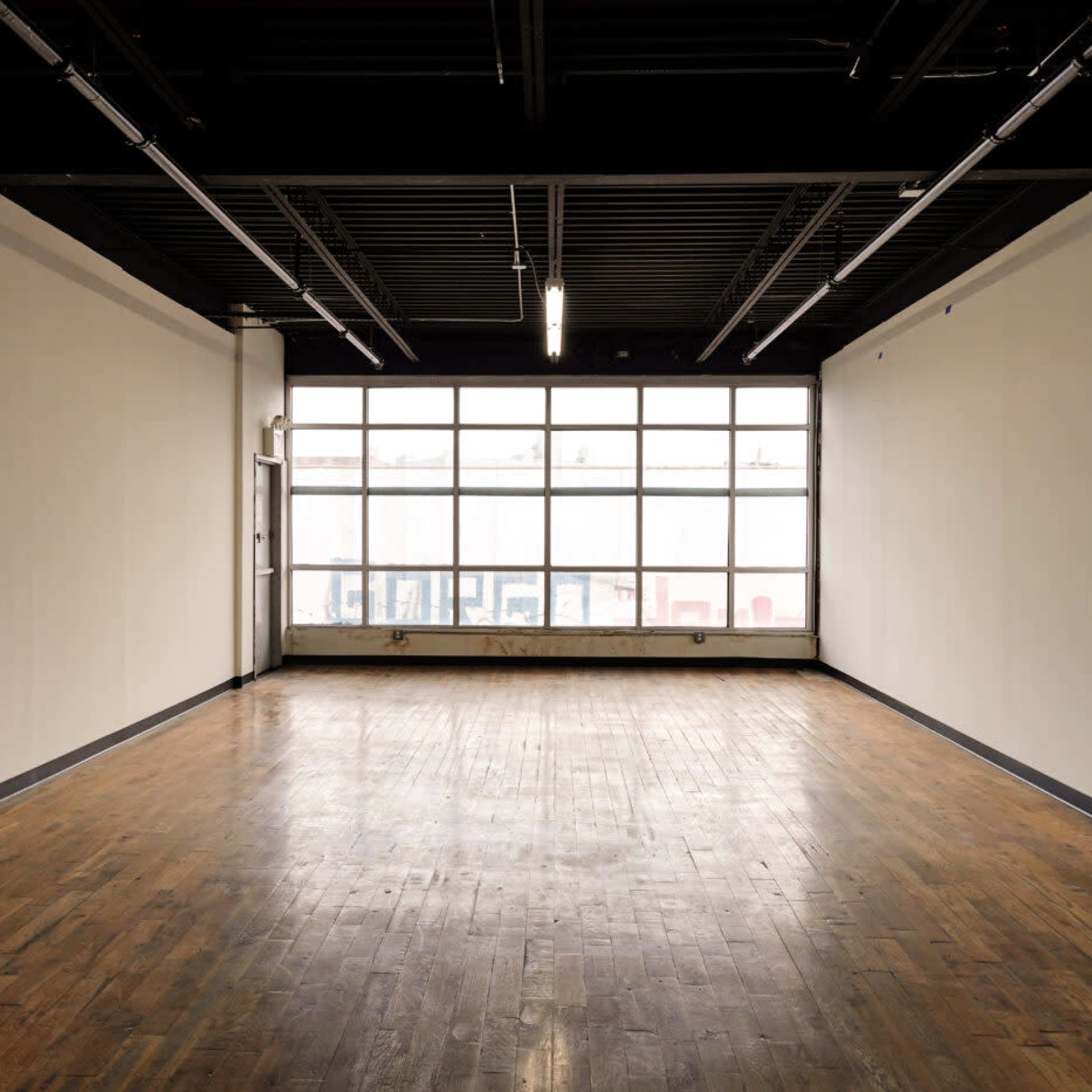 An empty room with wooden flooring and large windows along one wall, allowing natural light to enter the space.