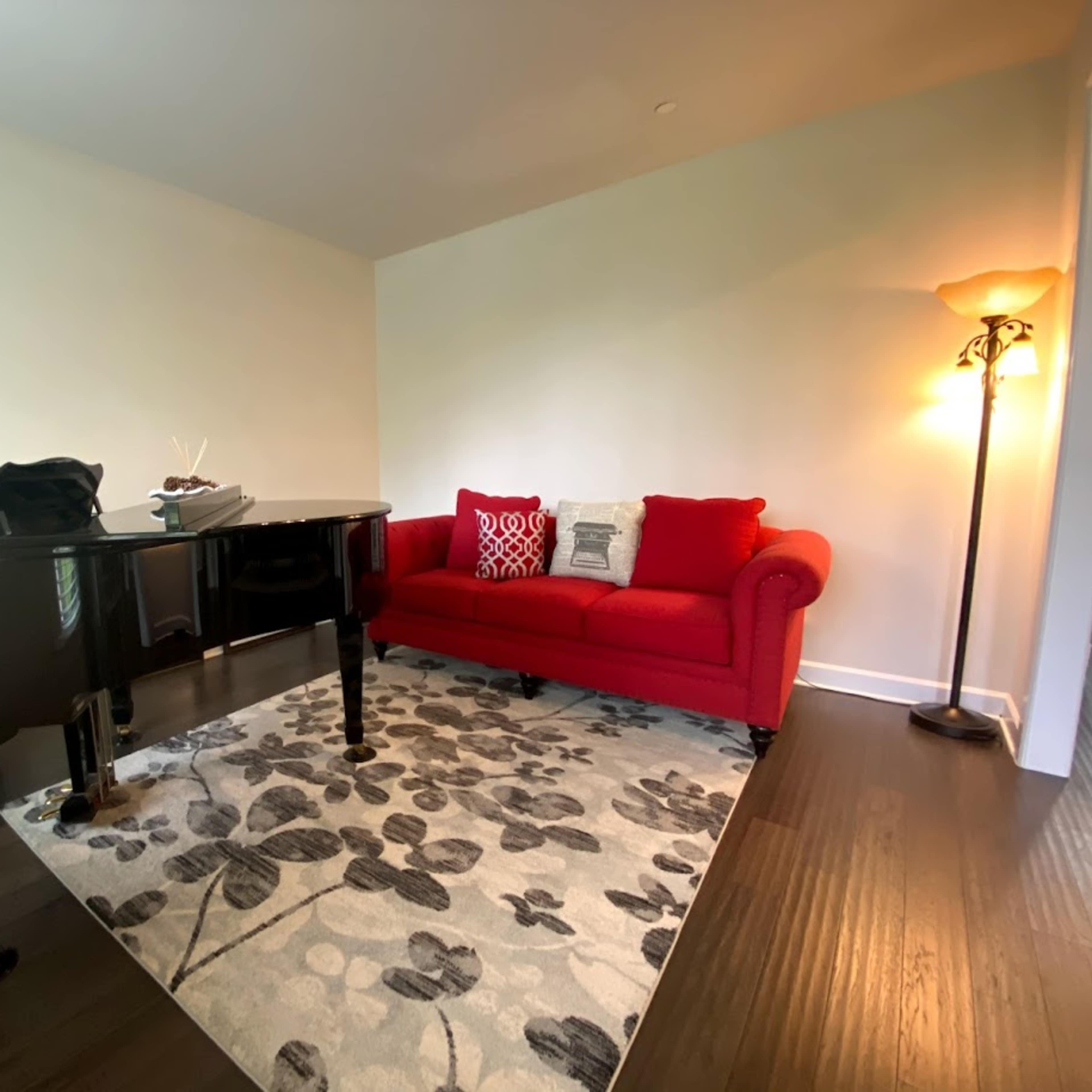A room with a black grand piano, a red sofa with decorative pillows, and a standing lamp on a floral rug.
