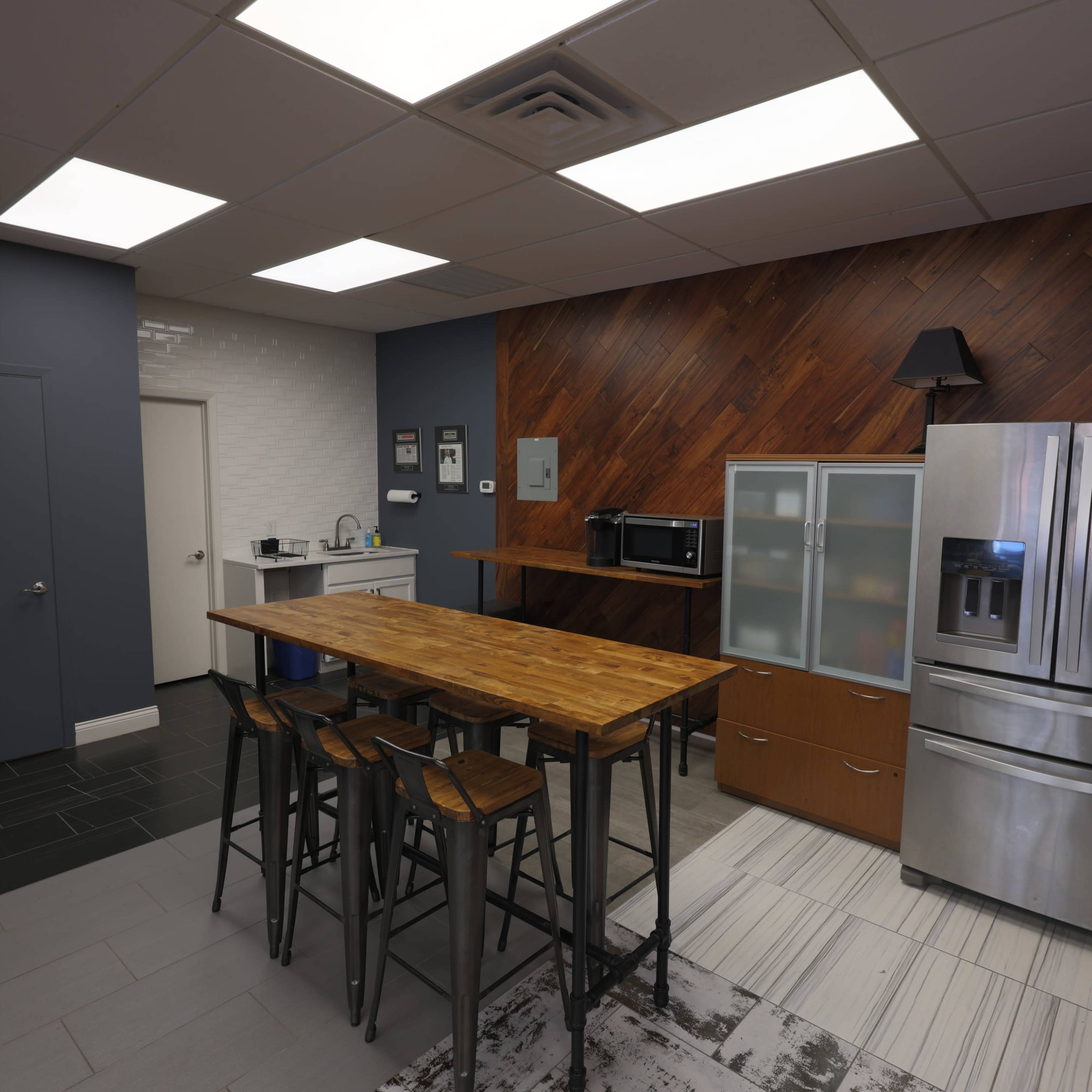 A modern kitchen with a wooden table and black stools, a stainless steel refrigerator, a microwave, and a glass-front cabinet against a wood-paneled wall.
