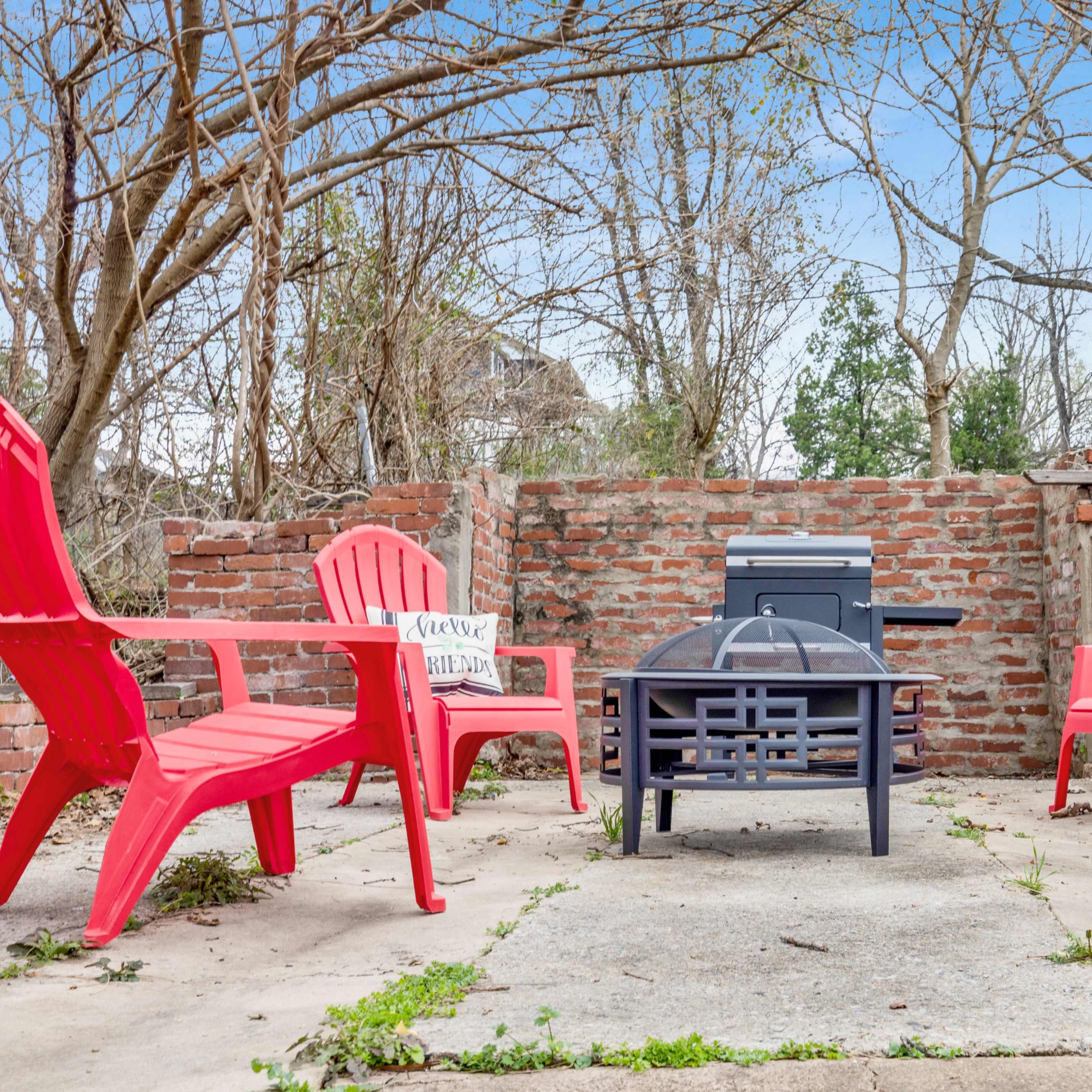 A small outdoor seating area features four red chairs arranged around a central concrete surface, with a grill and brick walls in the background.