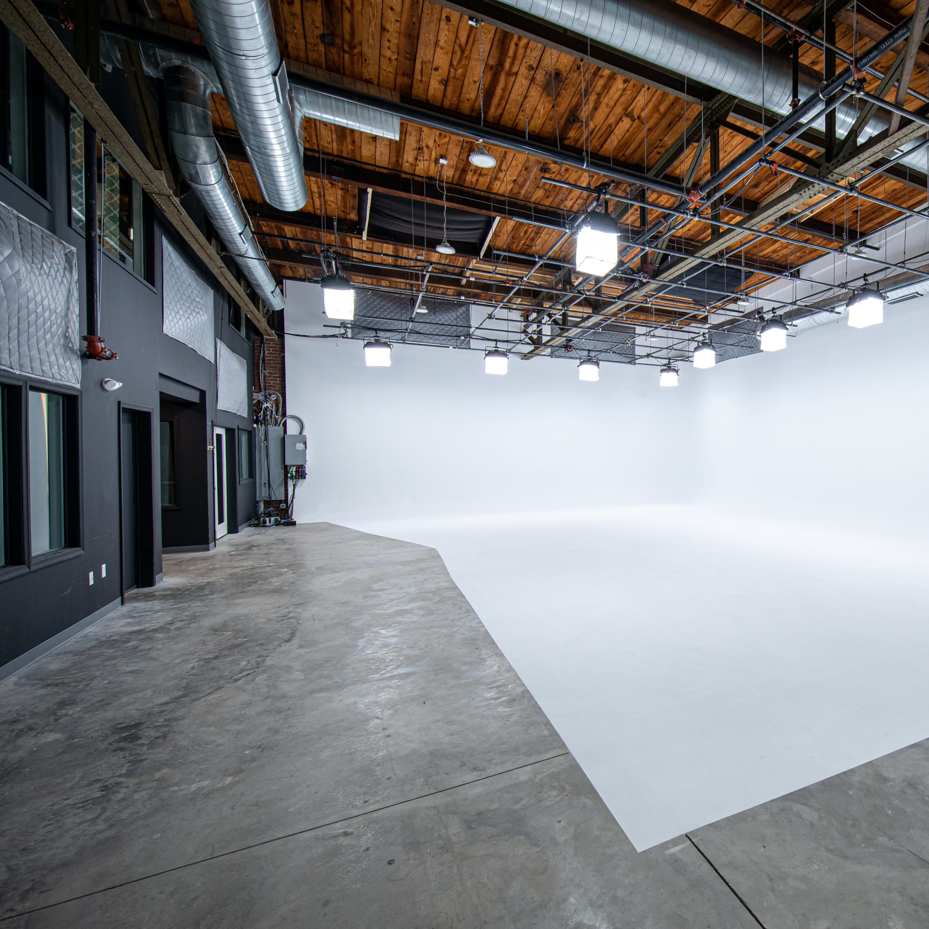 A spacious, empty studio with a white backdrop, wooden ceiling beams, and overhead lighting.