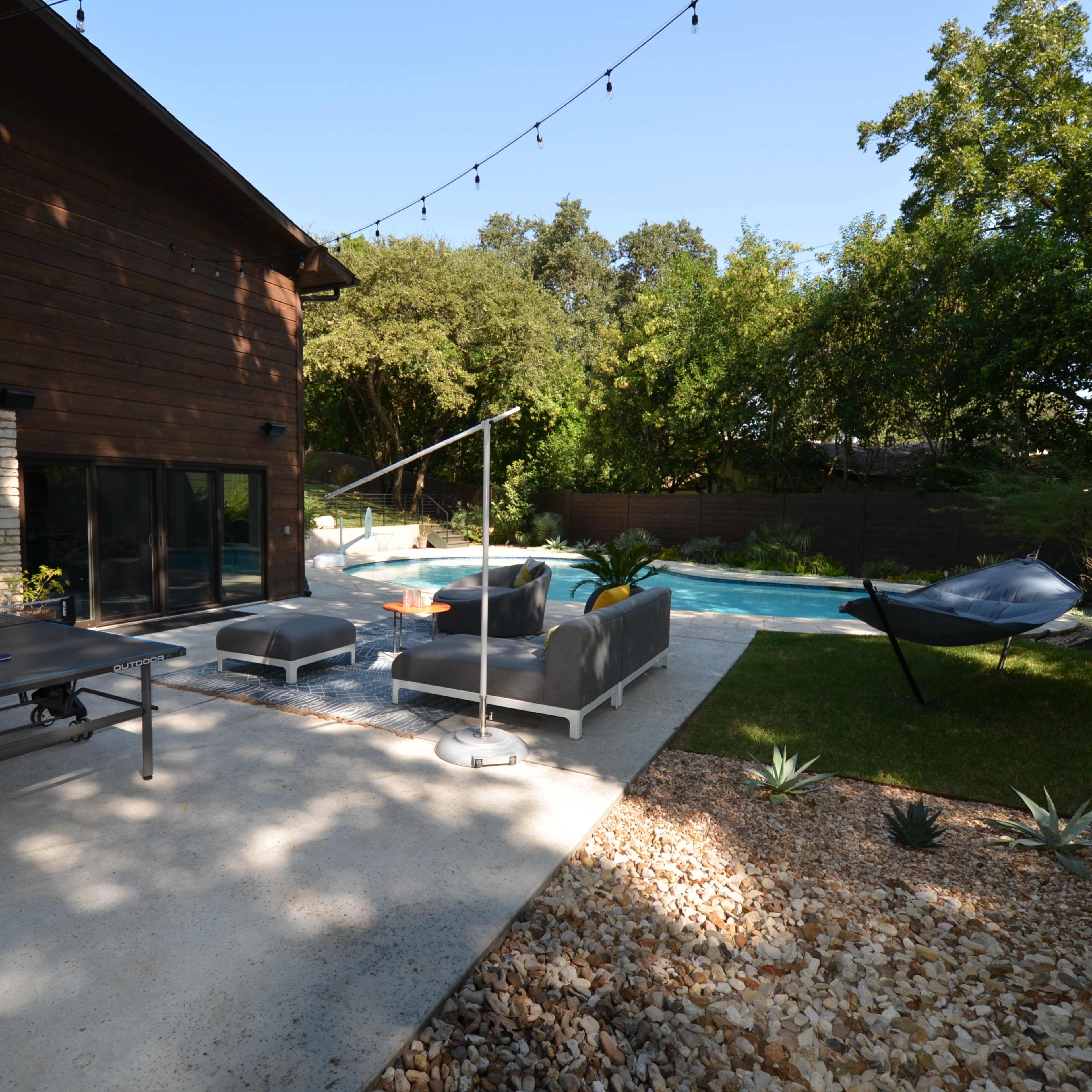 A backyard patio with a ping pong table, outdoor seating area, and a swimming pool surrounded by greenery.