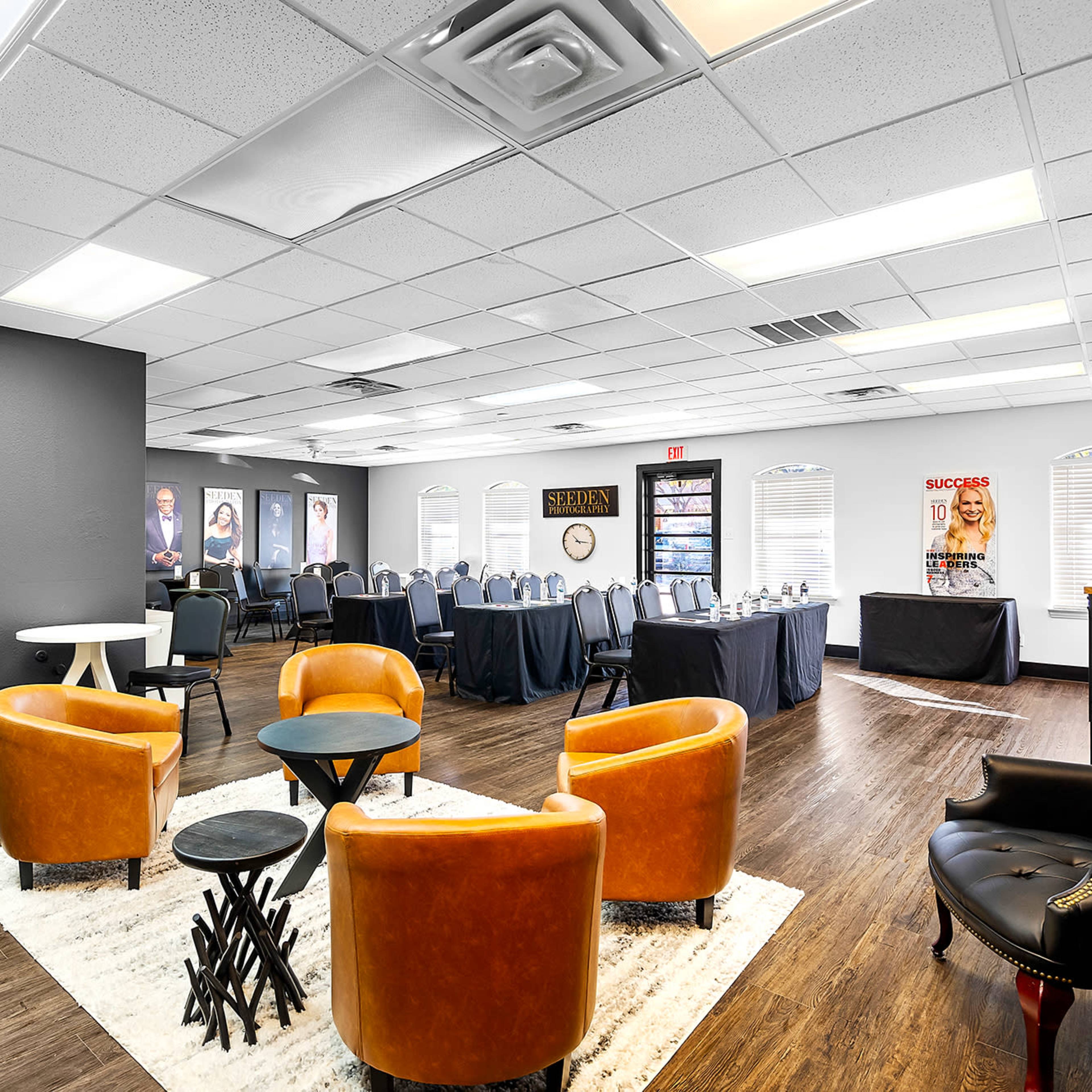A modern event space with a seating area featuring orange chairs and a table, alongside a setup for tables arranged for a gathering in the background.