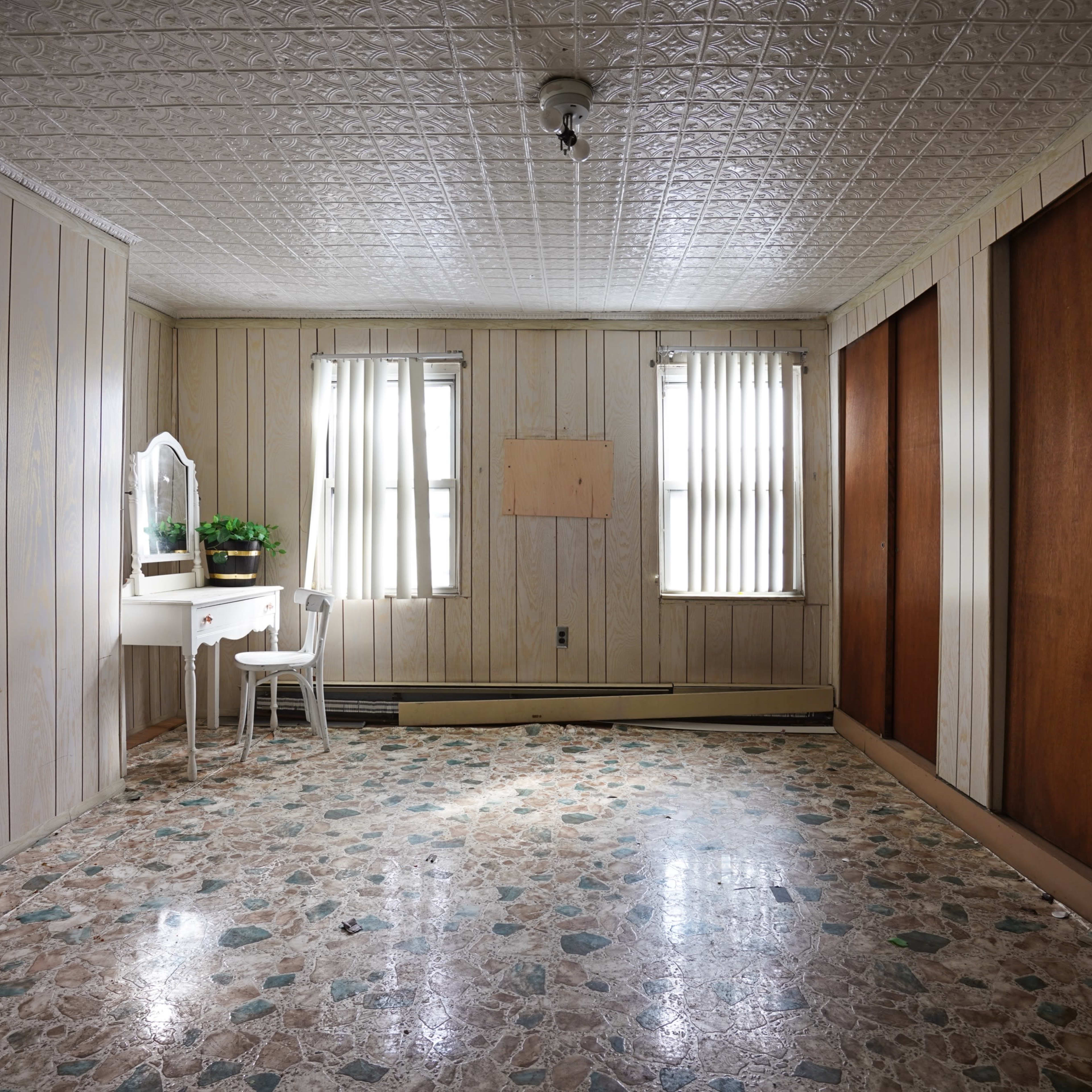 A sparsely furnished, empty room with wooden paneling, two windows with vertical blinds, and a small white vanity against one wall.