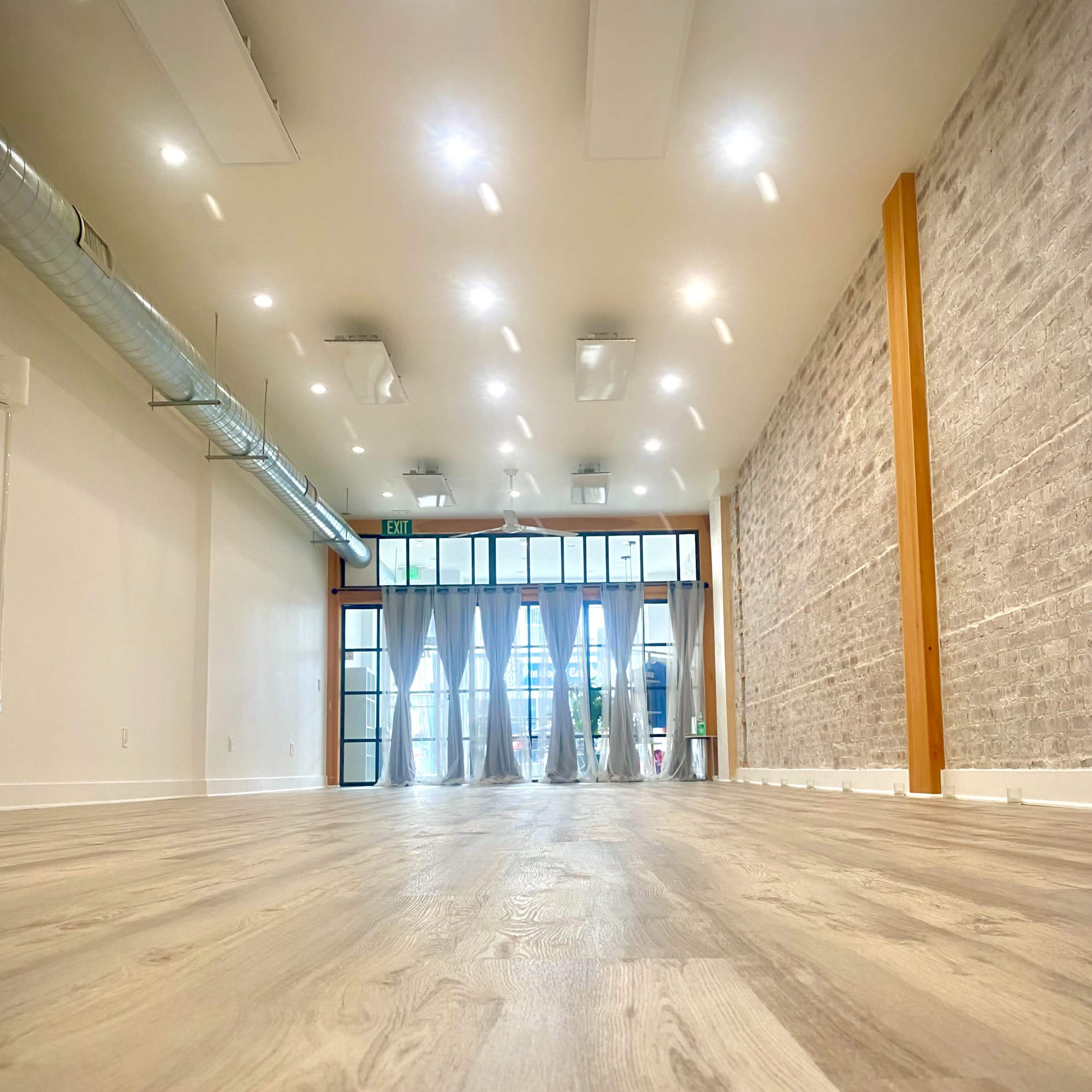 A spacious interior with light-colored wooden flooring, modern lighting fixtures, and large windows along one side, providing a view of the exterior.