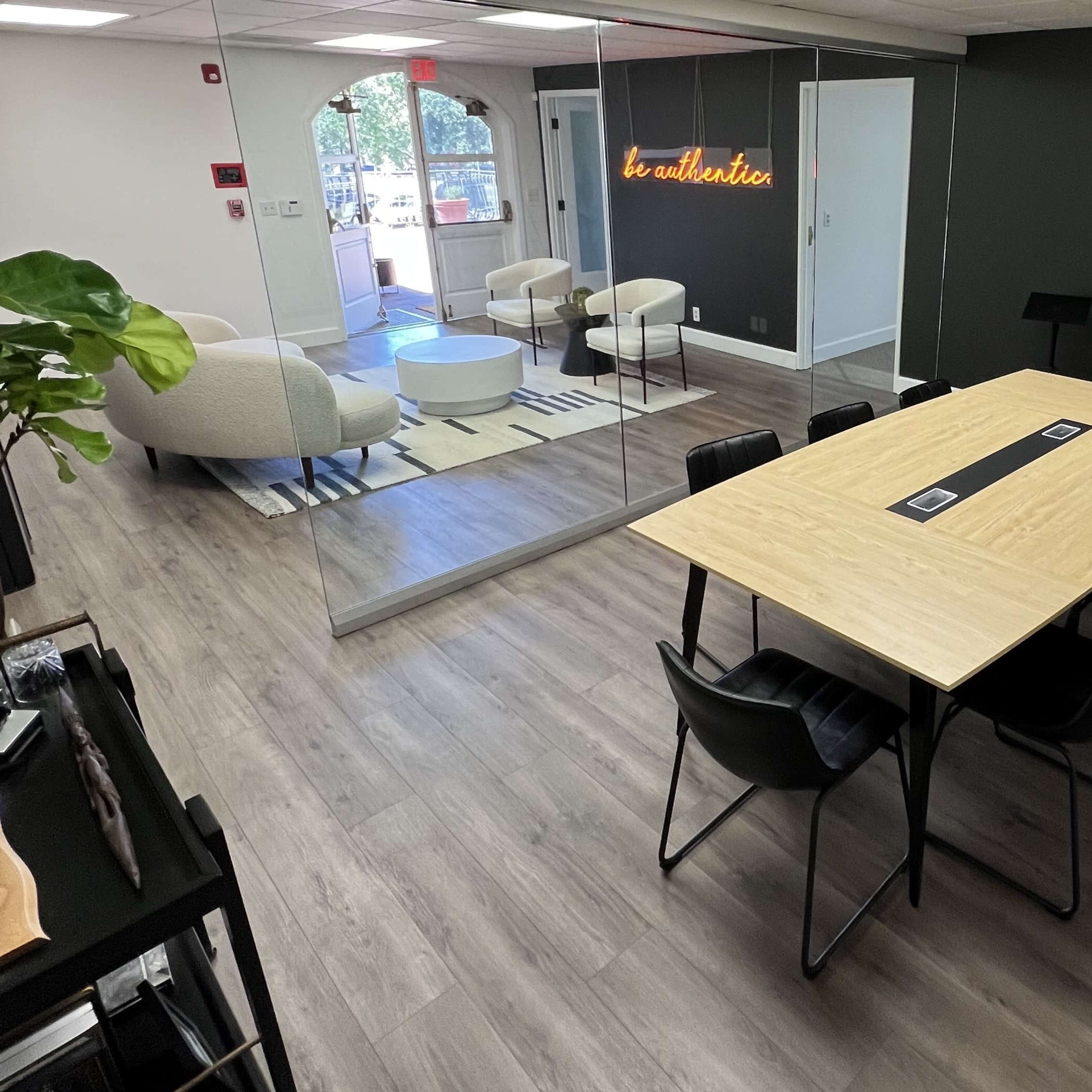 A modern office space featuring a large wooden conference table surrounded by black chairs, with a lounge area visible in the background.