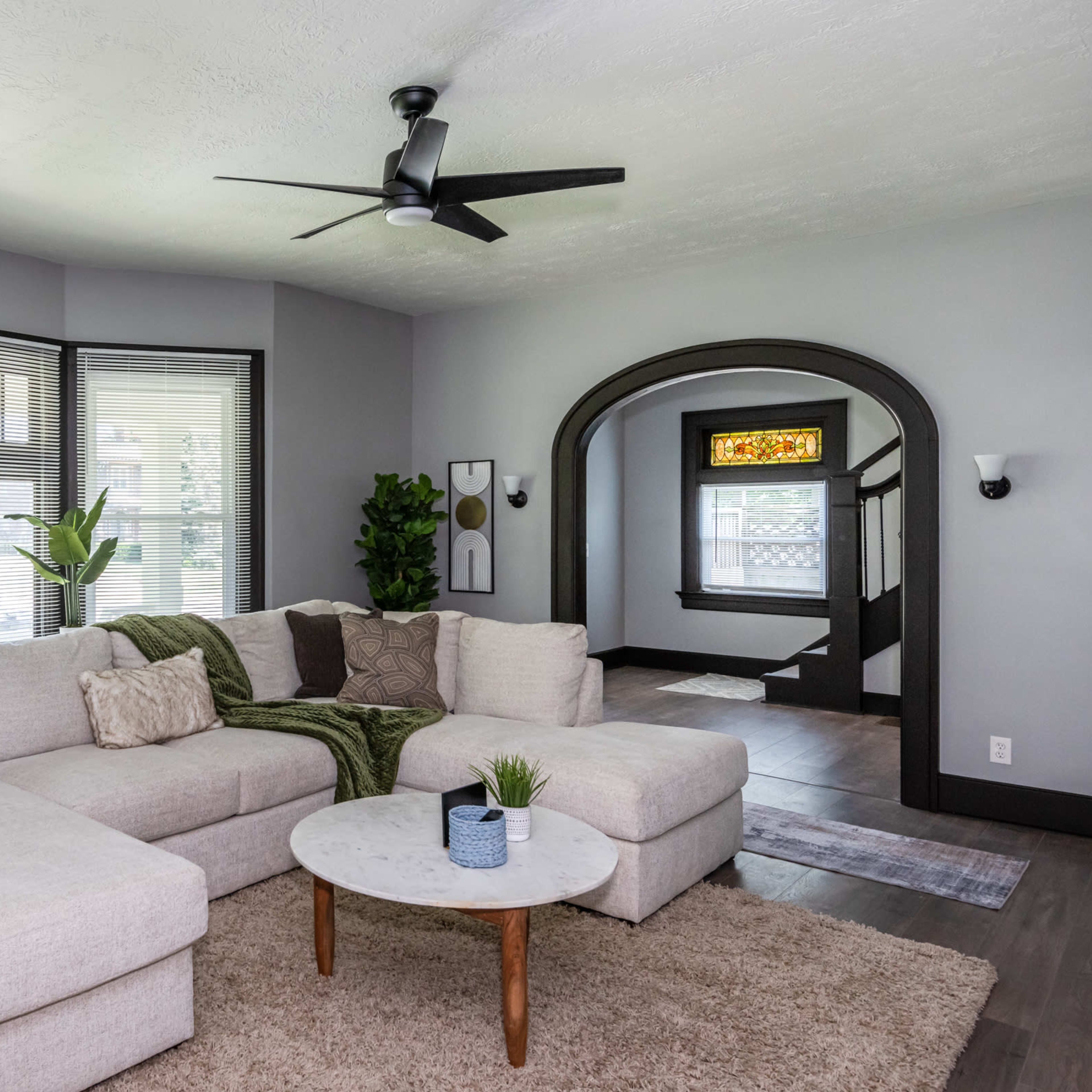 An open living room features a large sectional sofa, a round coffee table, and large windows with natural light.