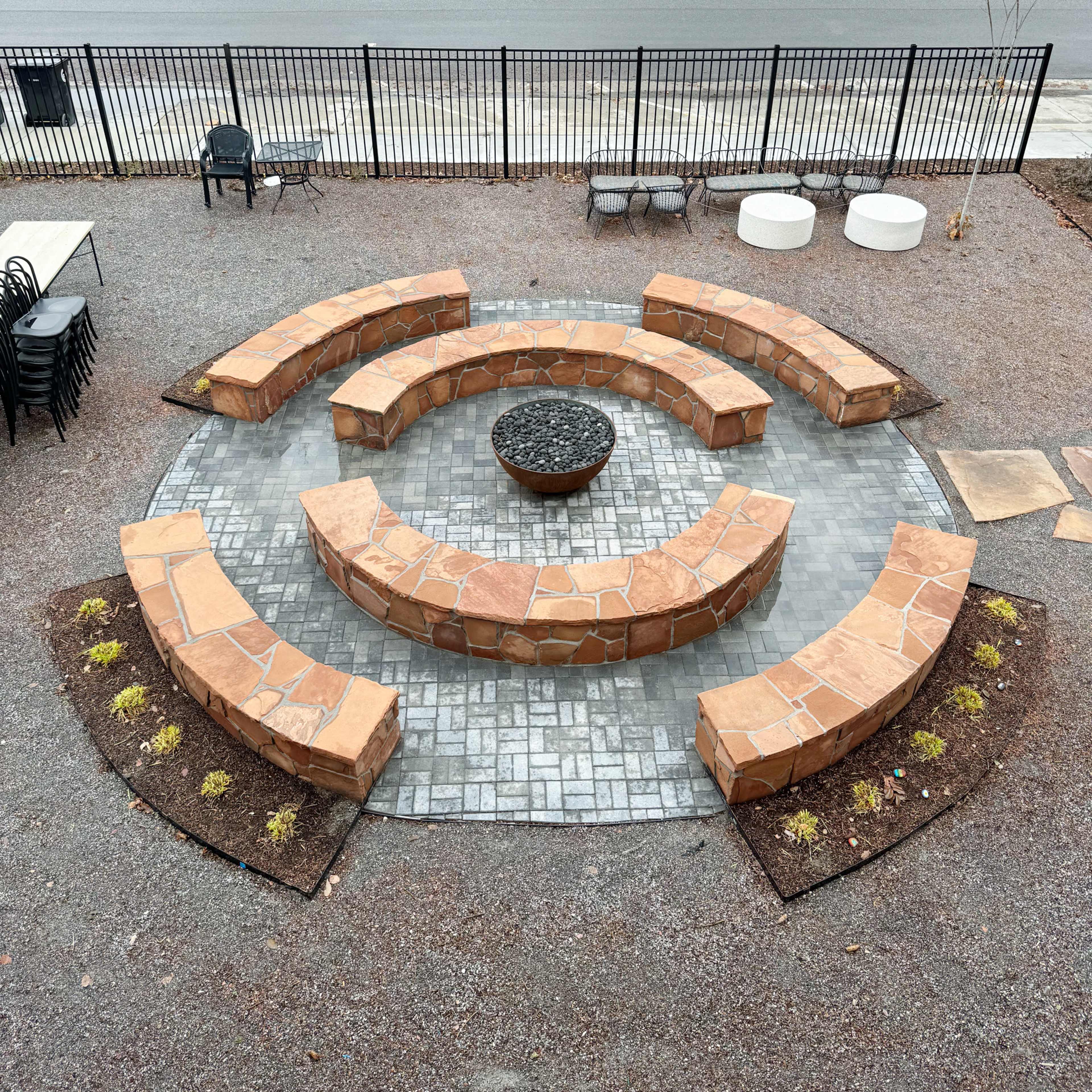 An outdoor circular seating area made of stone, with a central fire pit, surrounded by gravel and a fence.