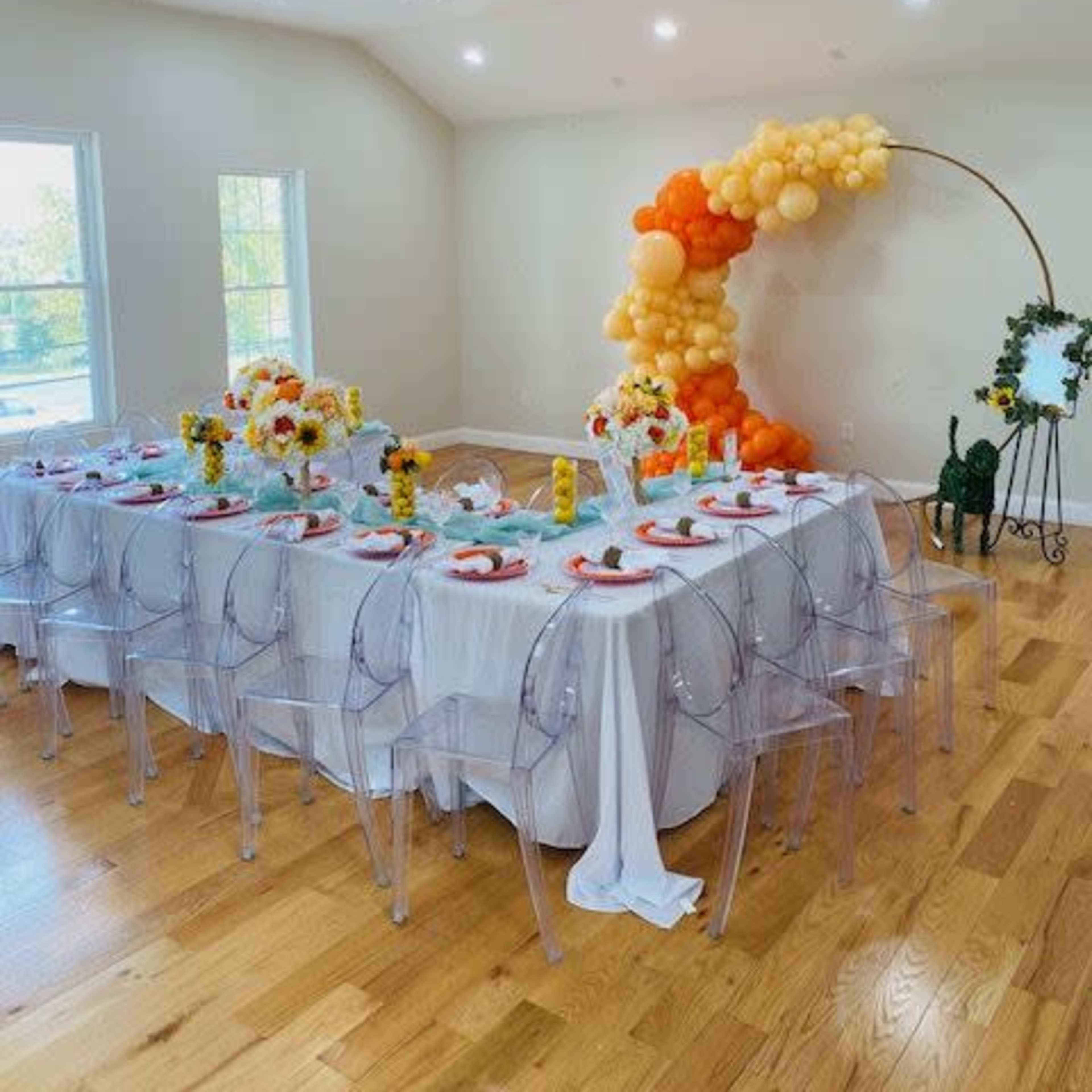 A long table set for a celebration features clear chairs and floral centerpieces, with a balloon arch in the background.