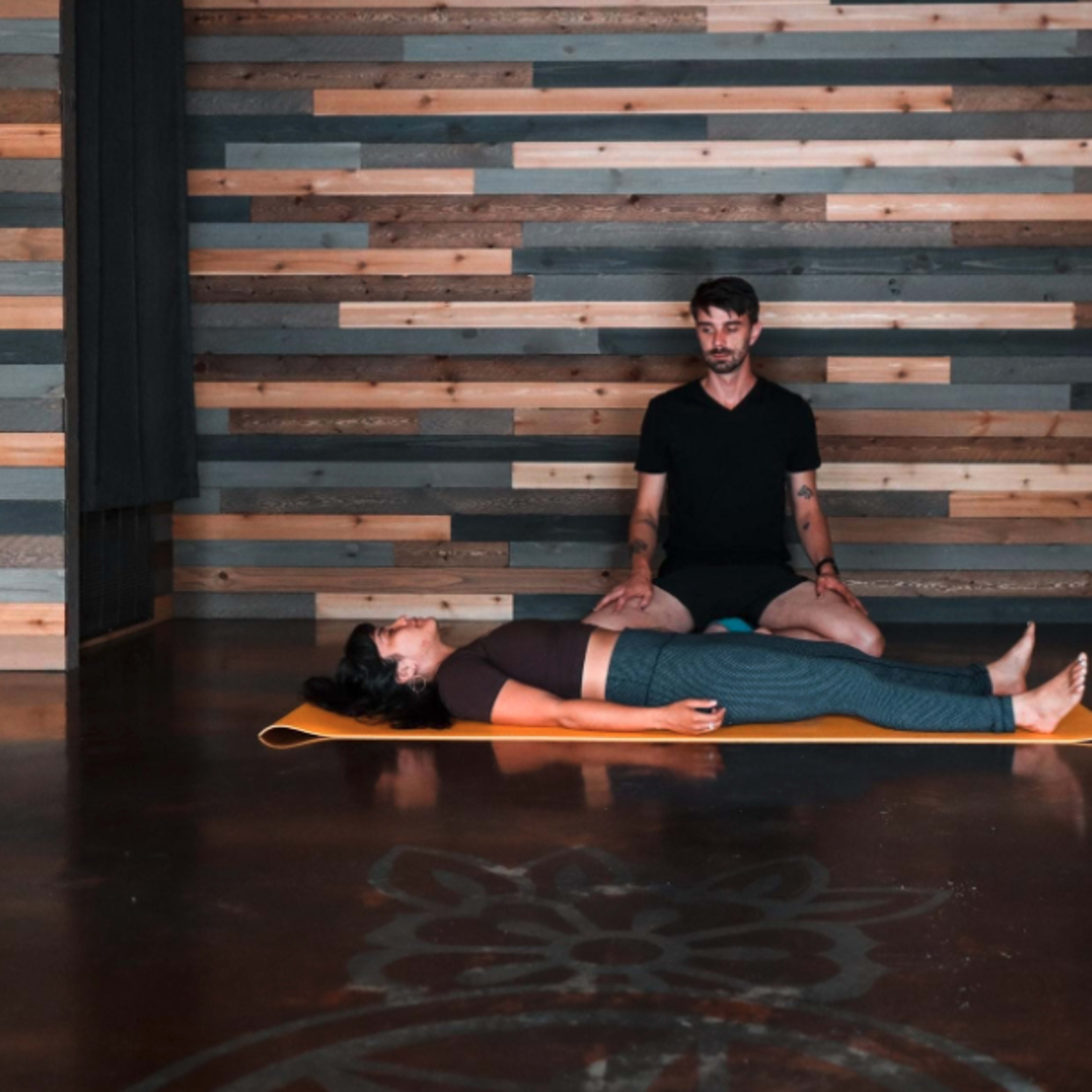 A person sits behind another person lying on a yoga mat in a studio with wooden walls.