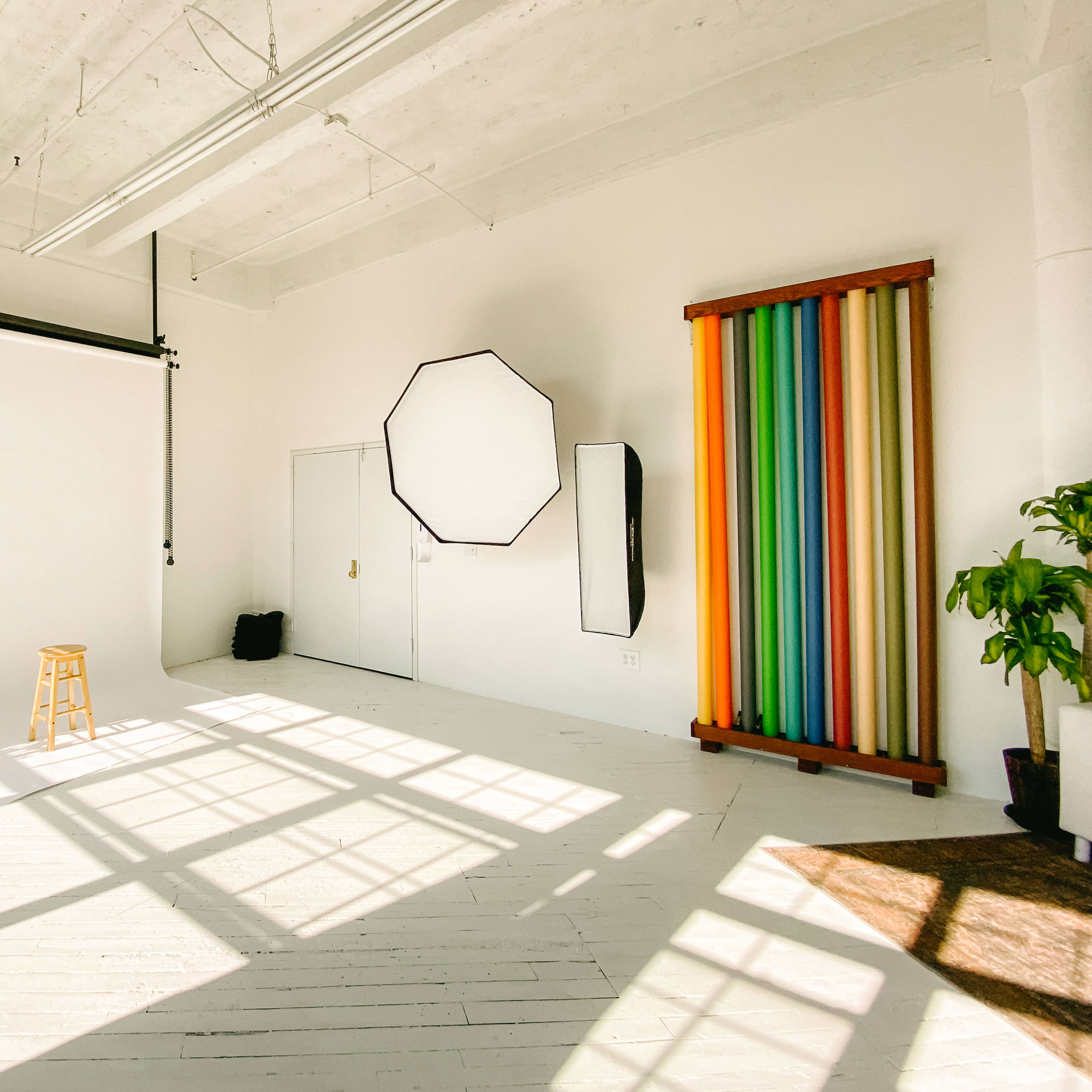 A bright photography studio with a wooden stool, a backdrop, and colorful fabric drapes hanging on the wall.