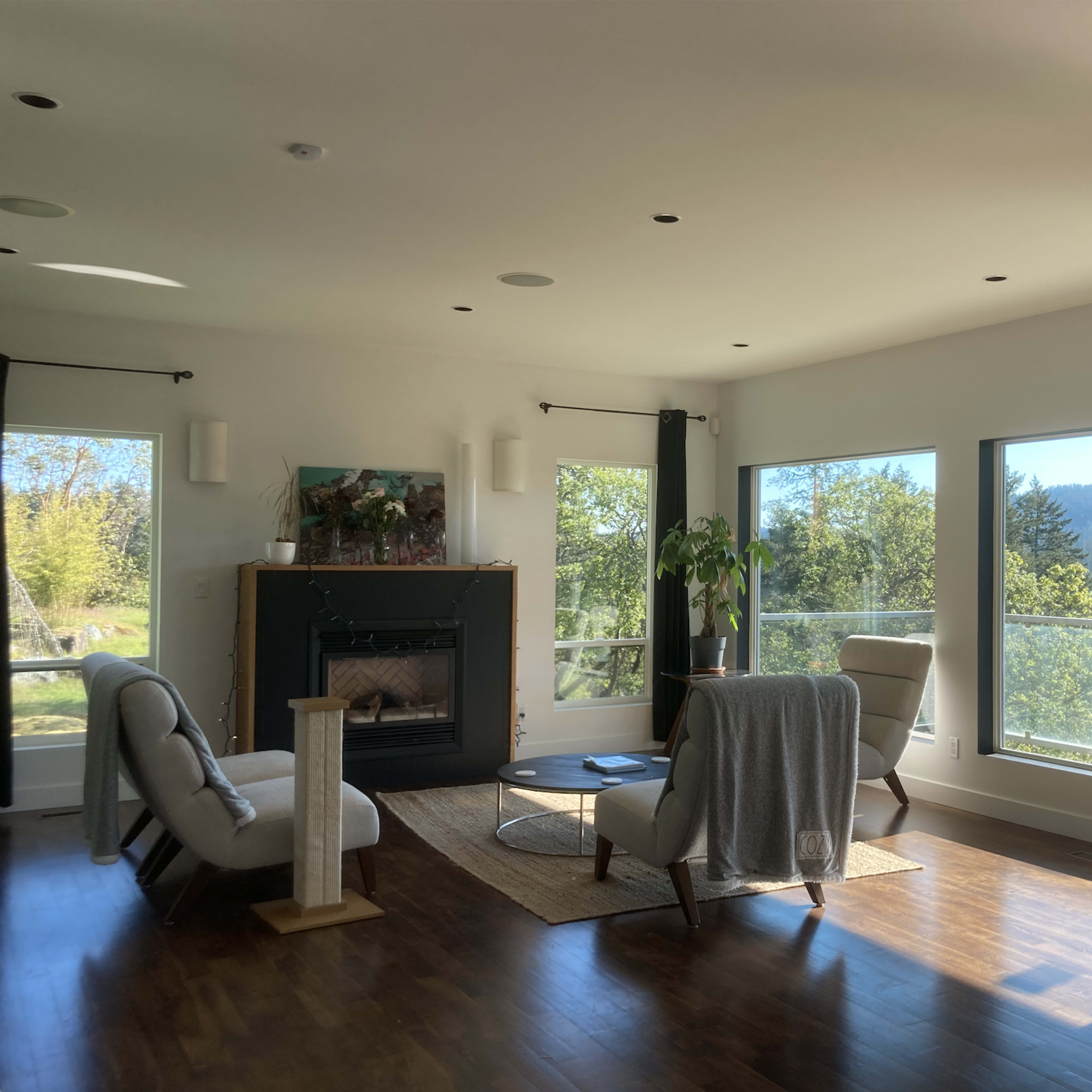 A modern living room features two chairs, a coffee table, and a fireplace, with large windows providing a view of the outdoors.