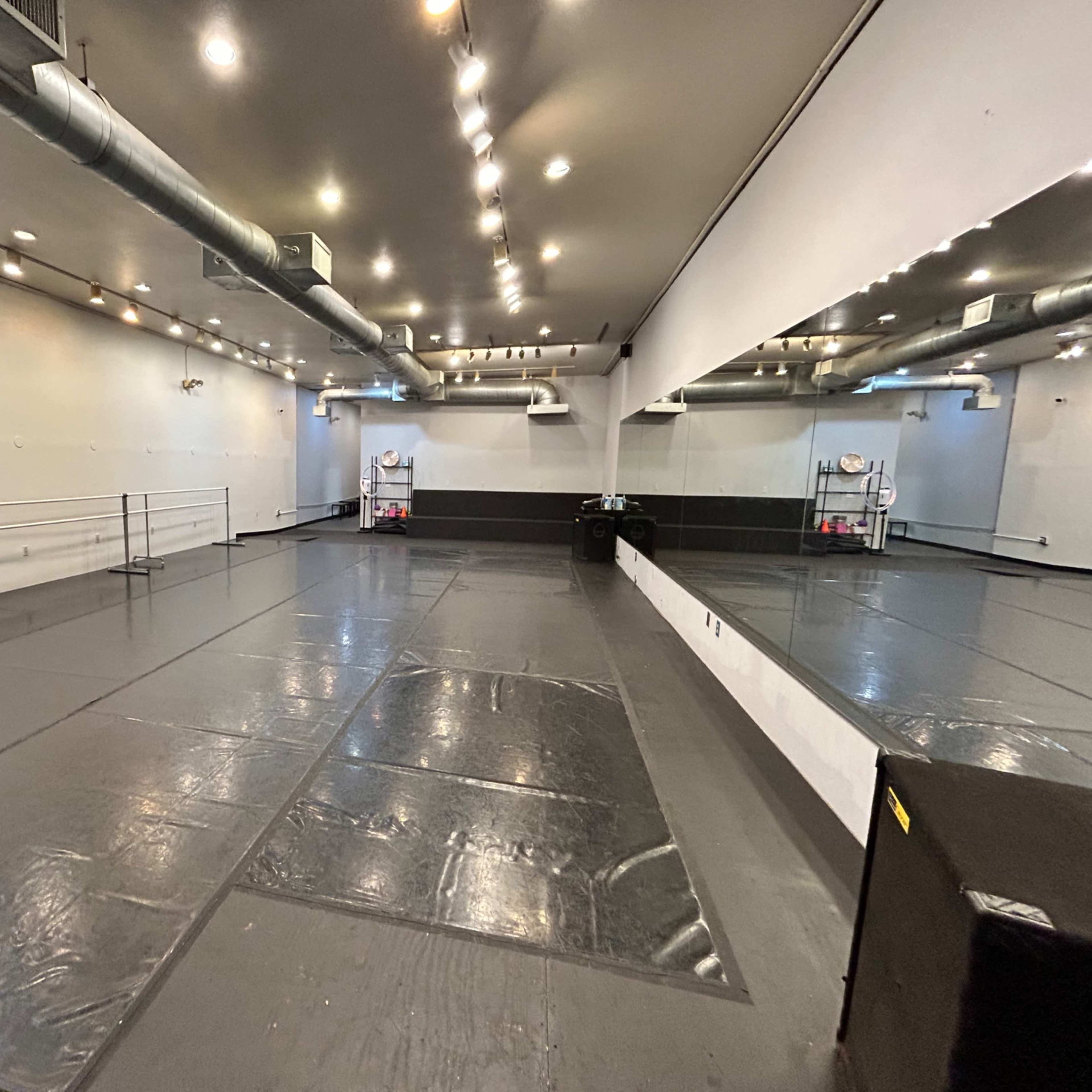 A spacious dance studio with a mirrored wall, dance barre, and grey flooring.