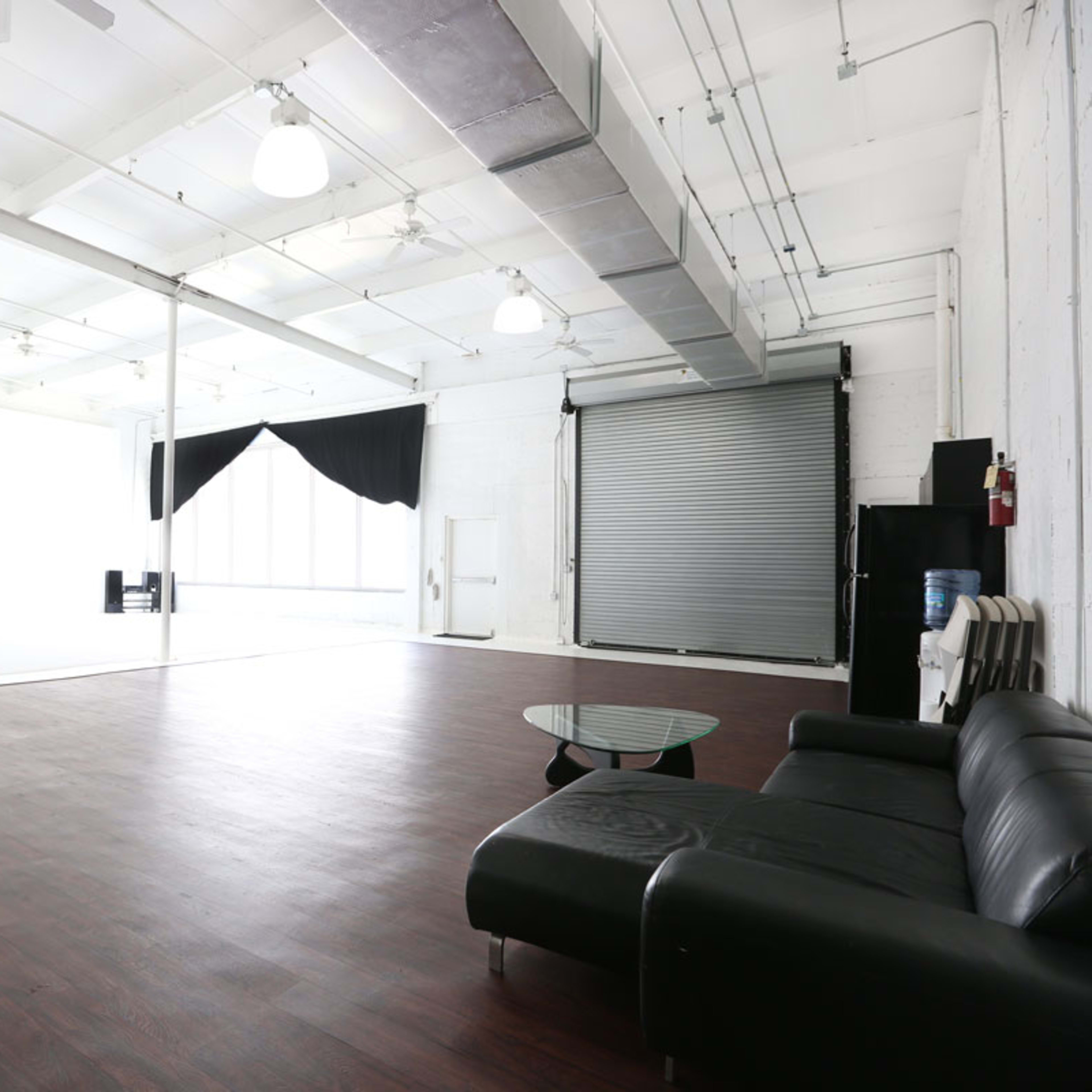 A spacious, empty studio with white walls, a dark wooden floor, a black couch, and large windows covered with sheer curtains.