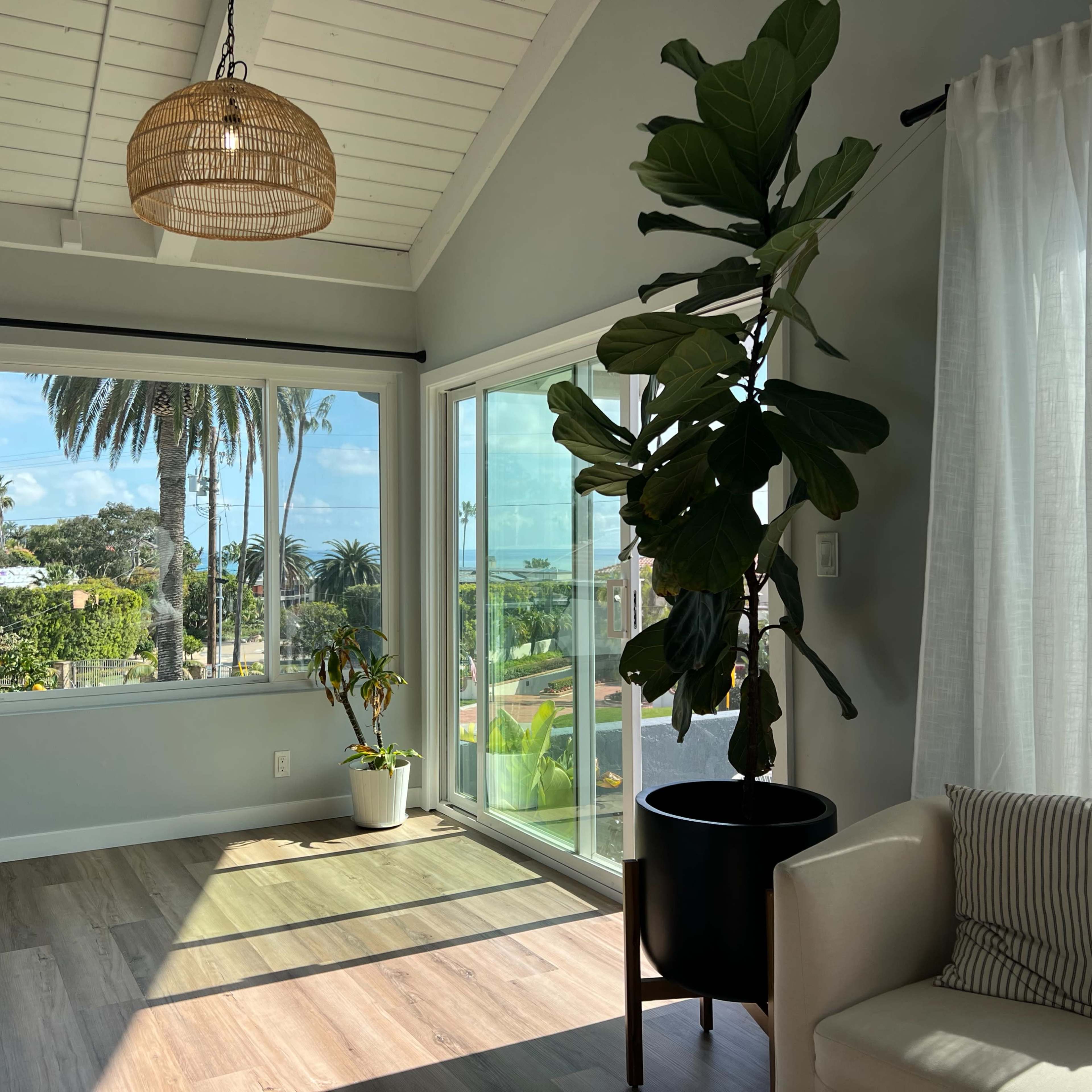 A well-lit room features large windows with a view of palm trees and a waterfront, along with a potted plant and a cozy seating area.