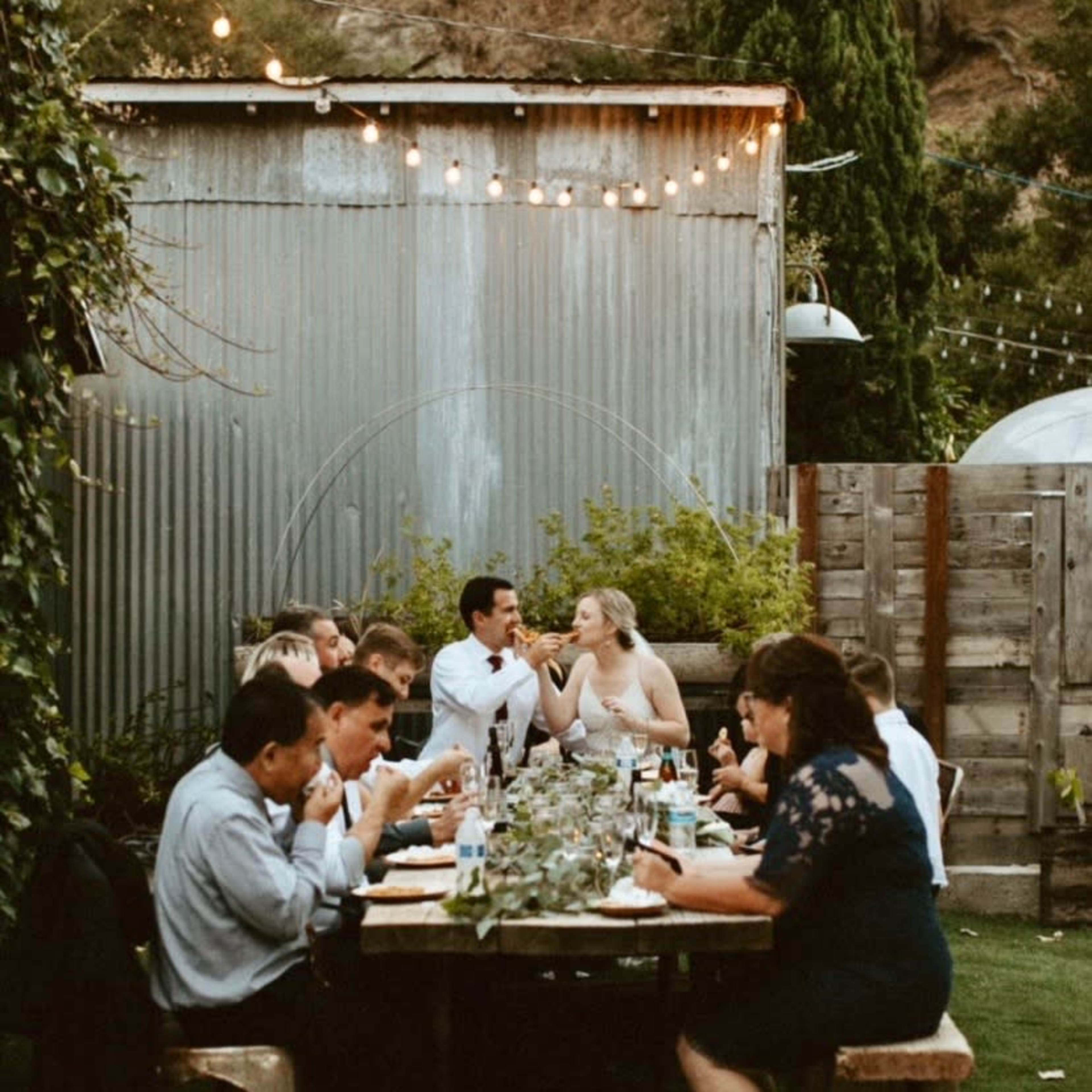 A group of people is gathered around tables for an outdoor dinner party, with string lights illuminating the setting and a rustic metal building in the background.