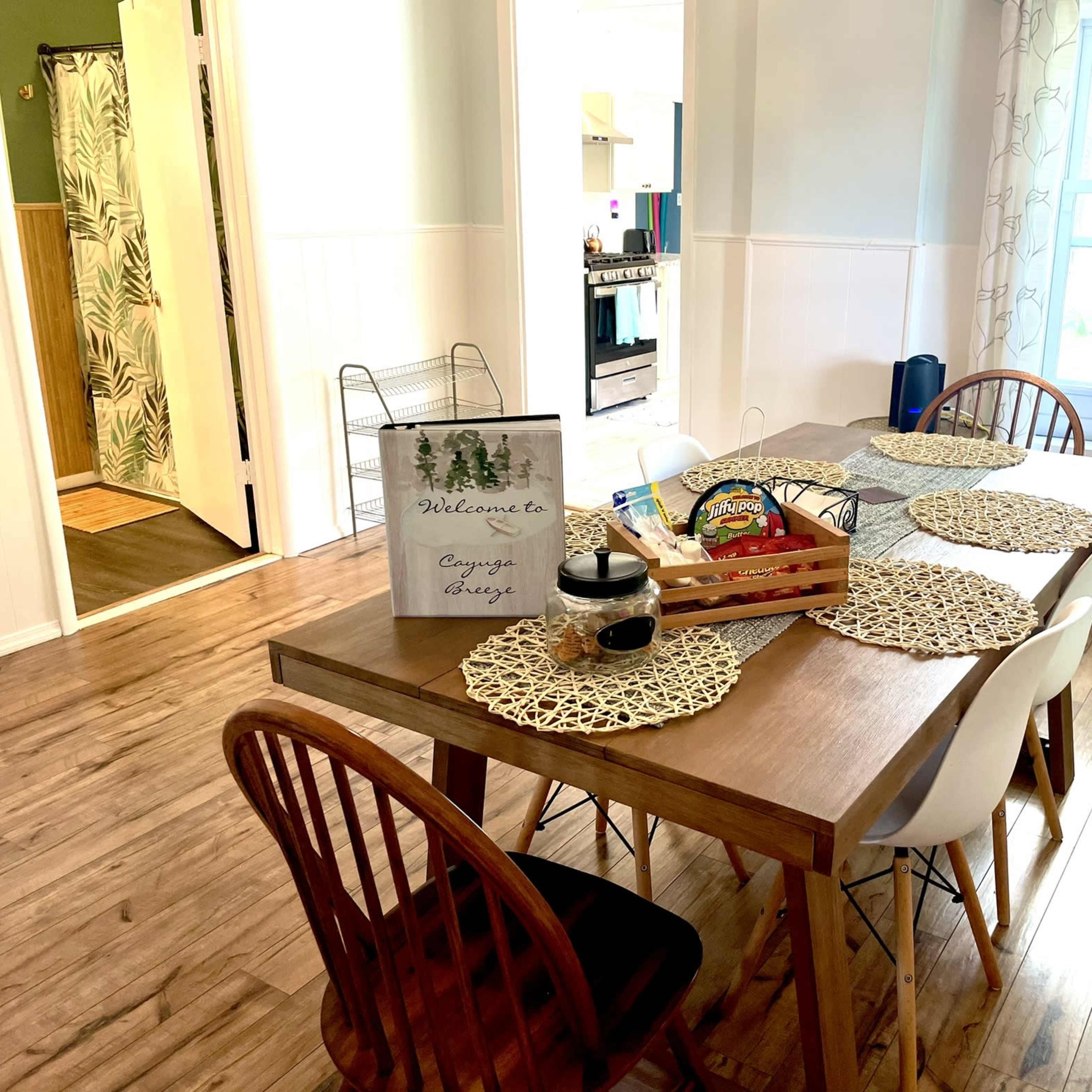 A dining table with placemats and a welcome sign is set in a well-lit room with a kitchen doorway and a fan visible in the background.