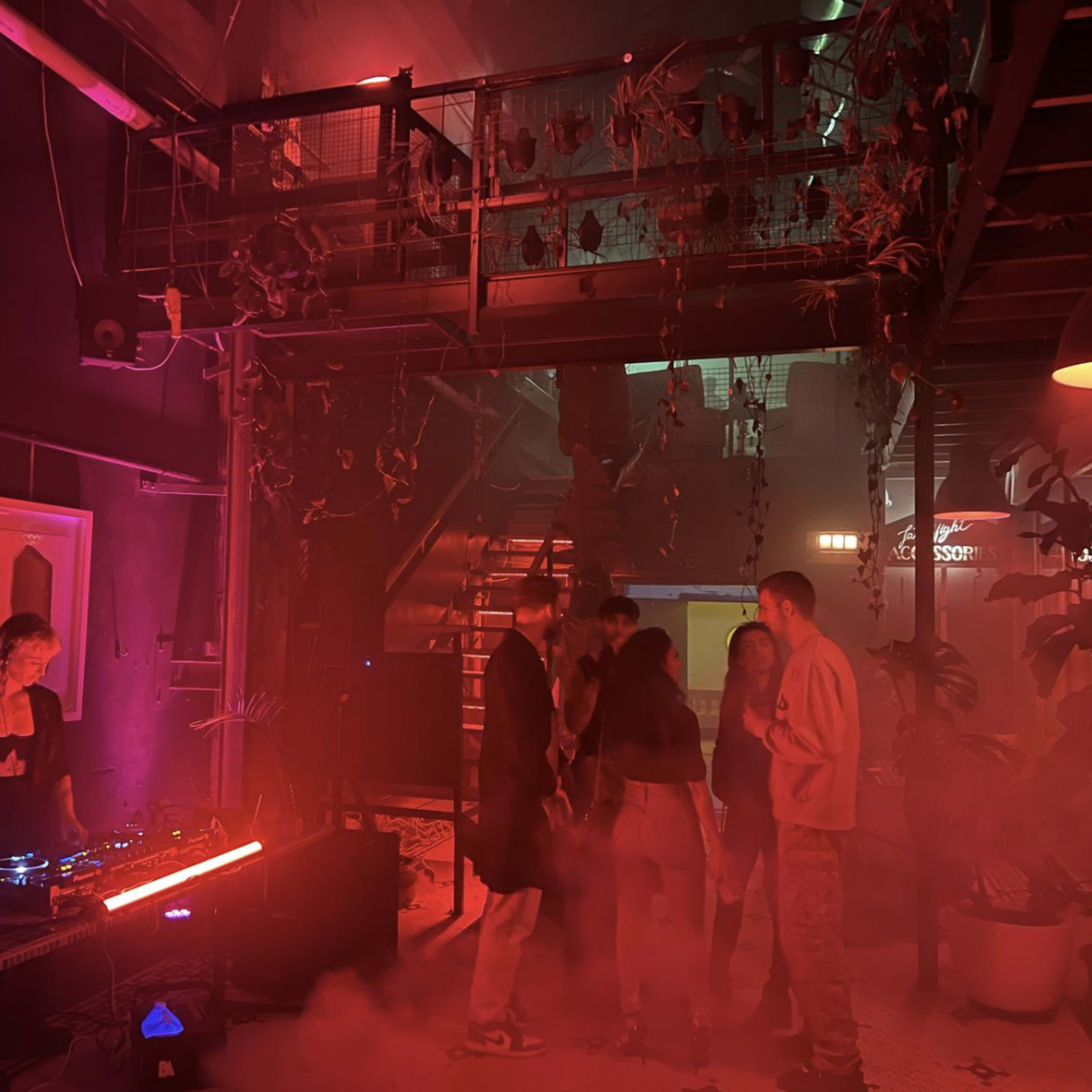 A DJ performs in a dimly lit venue filled with colorful lights and smoke while a small group of people dance nearby.