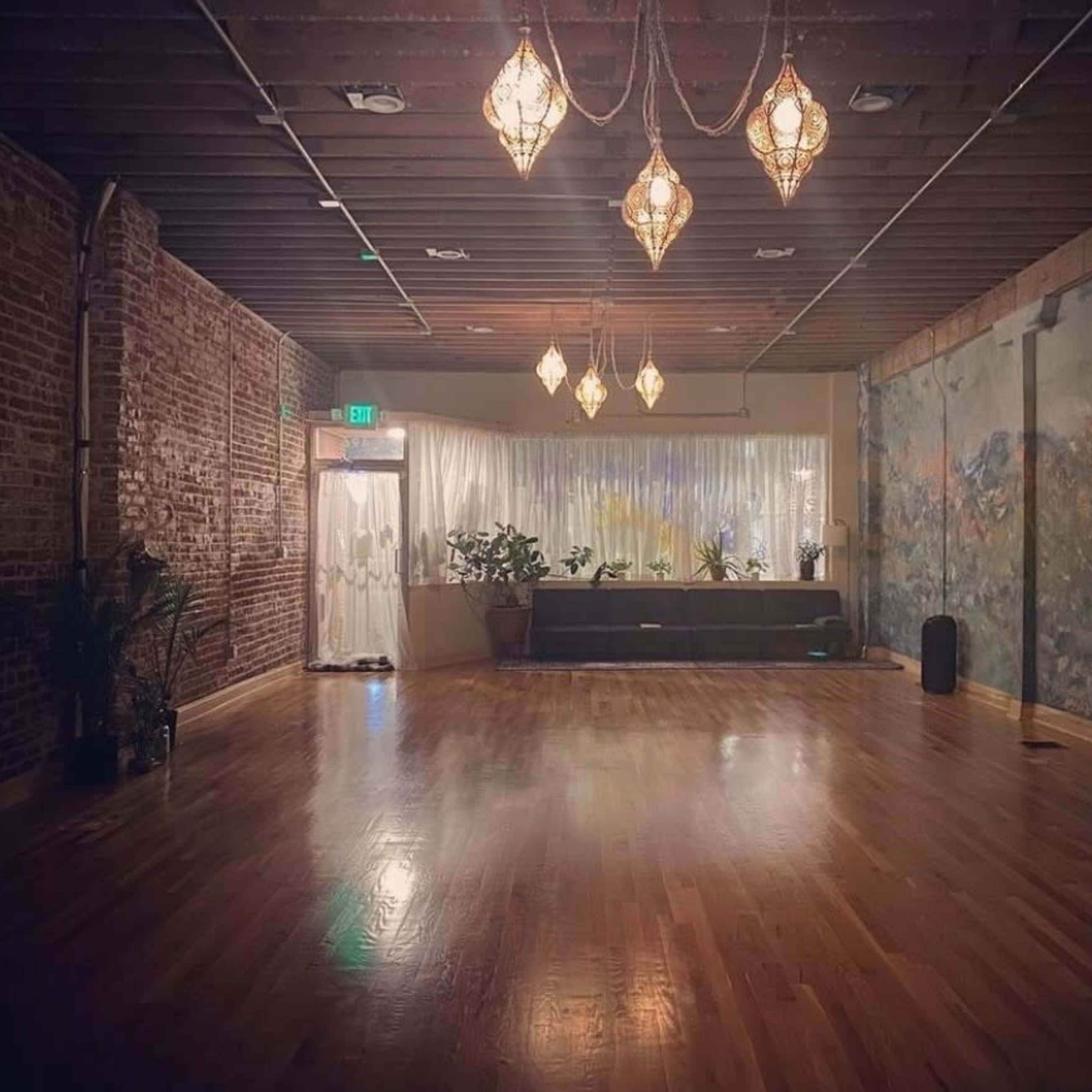 A spacious, well-lit room features hardwood floors, exposed brick walls, and decorative hanging lights, with a mural and plants along one side.