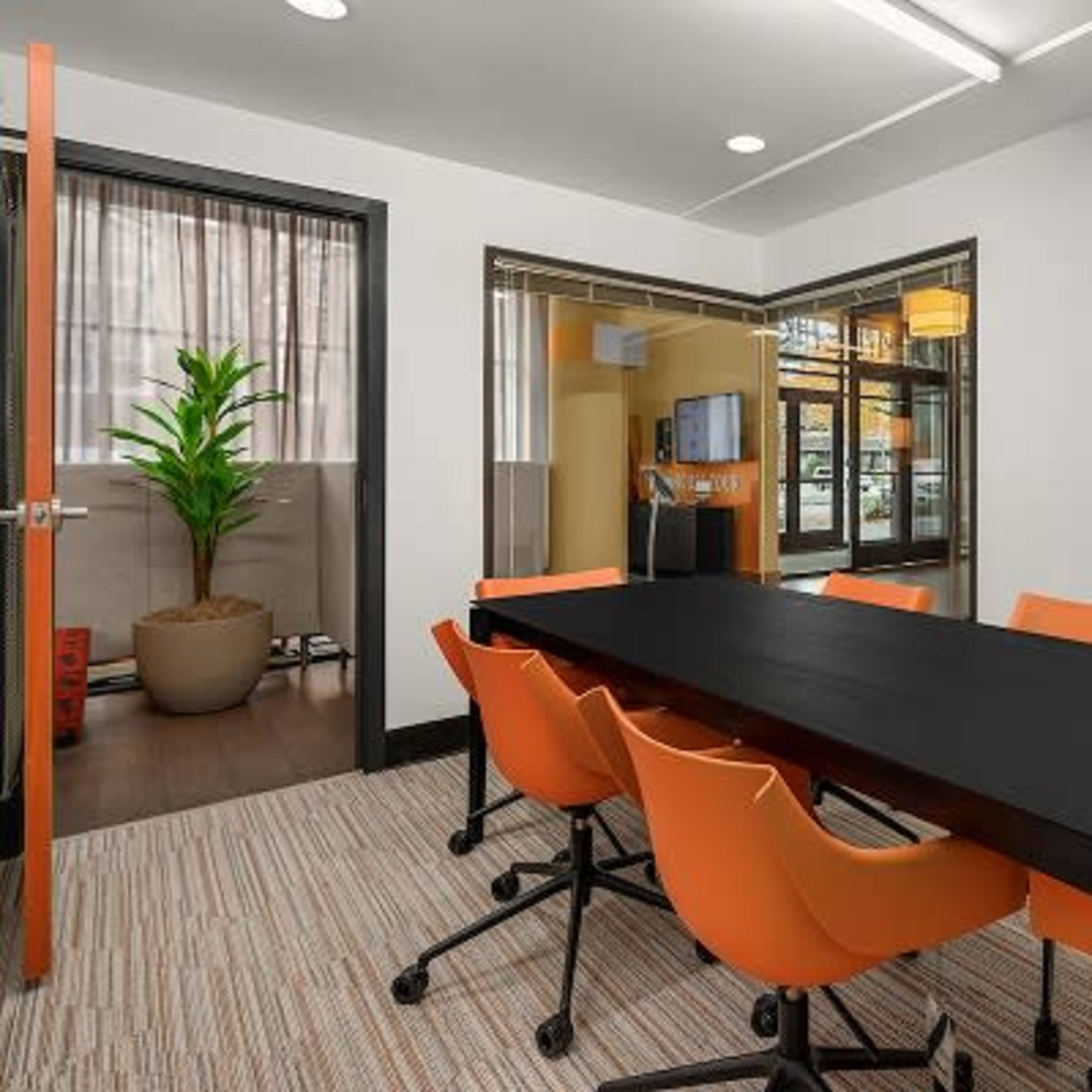 A conference room features a black table surrounded by six orange chairs, with a plant in a pot visible in the background.