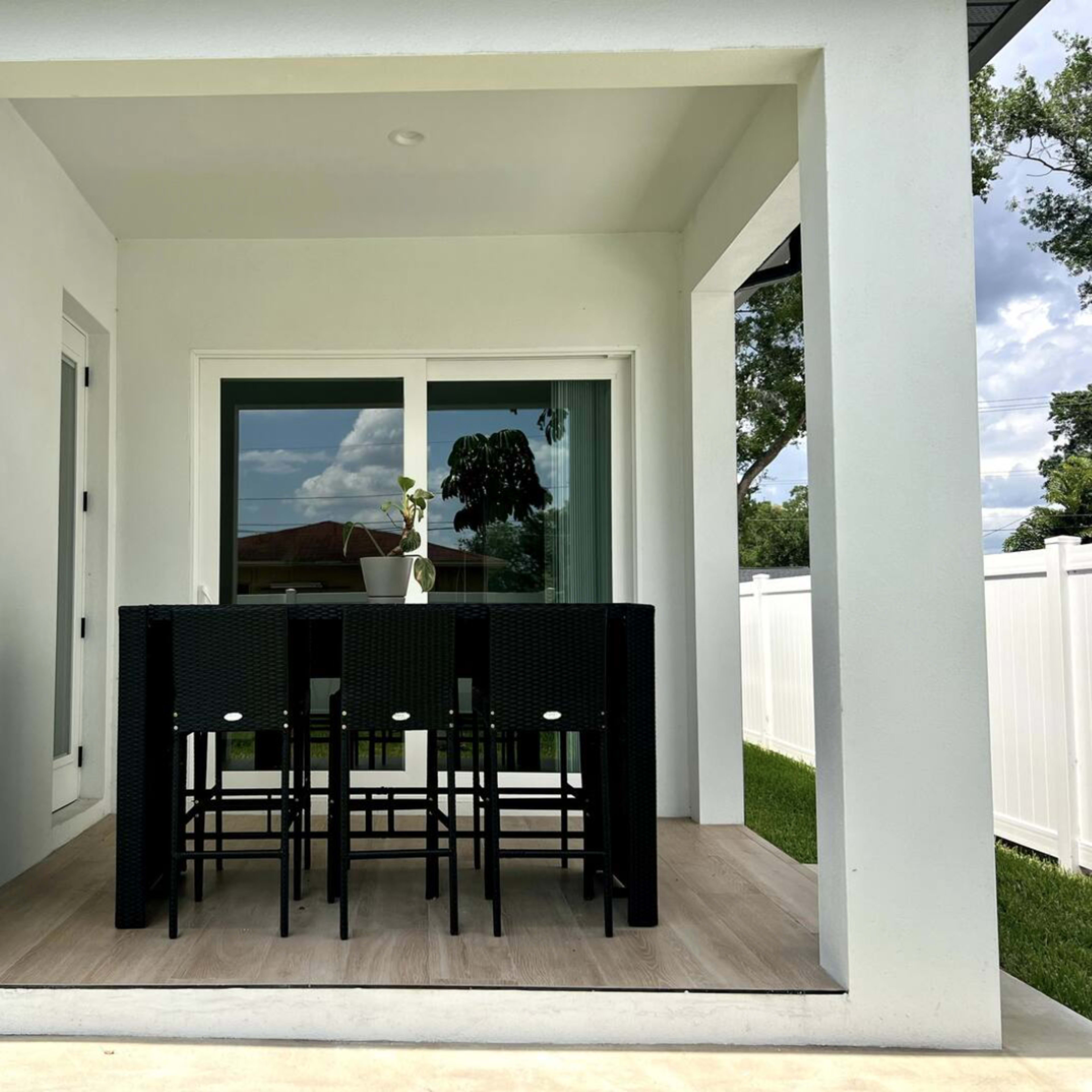 A covered patio features a black dining table with six chairs and a potted plant on the table, framed by white walls and a grassy area.