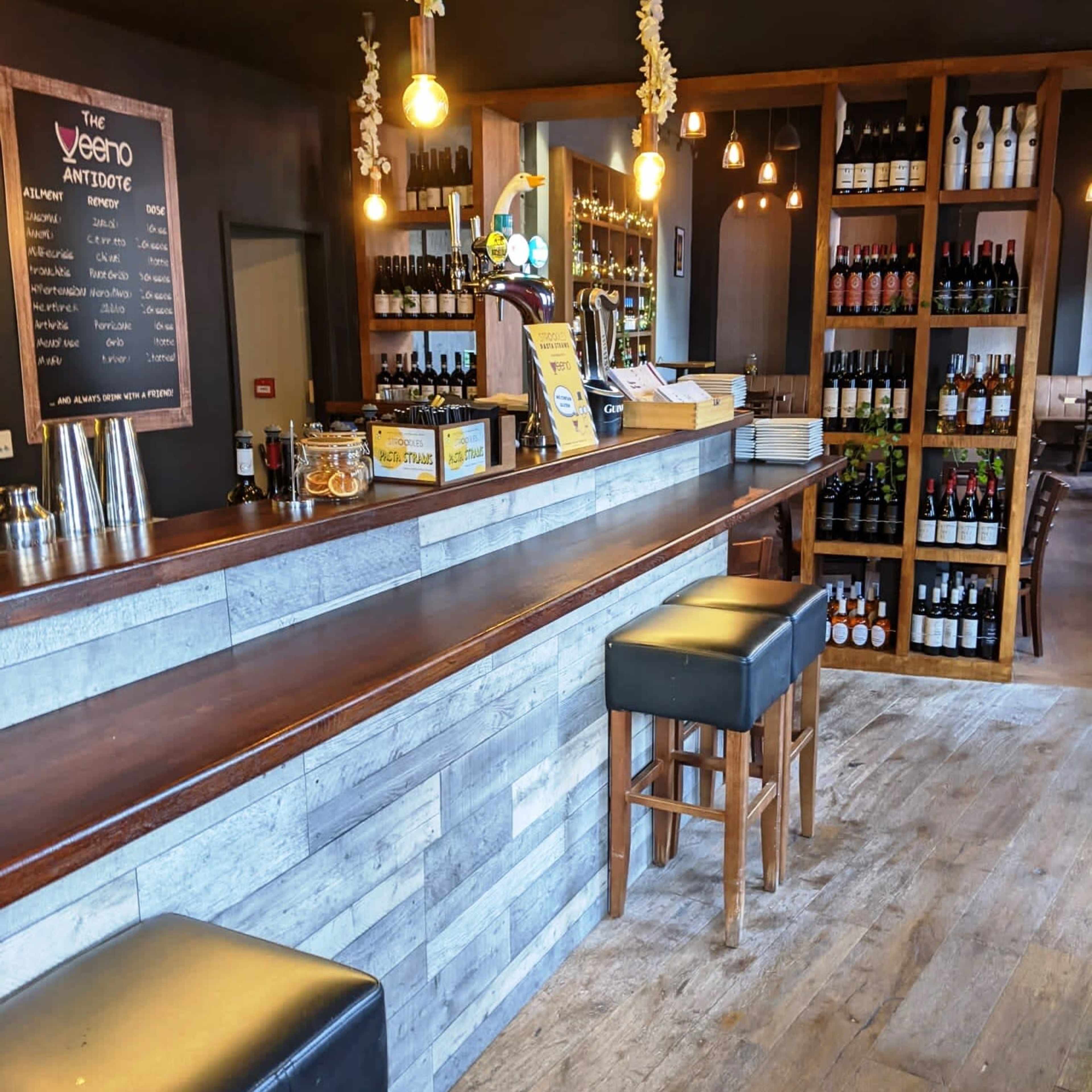 A bar area with a wooden counter, high stools, shelves filled with bottles, and warm lighting.