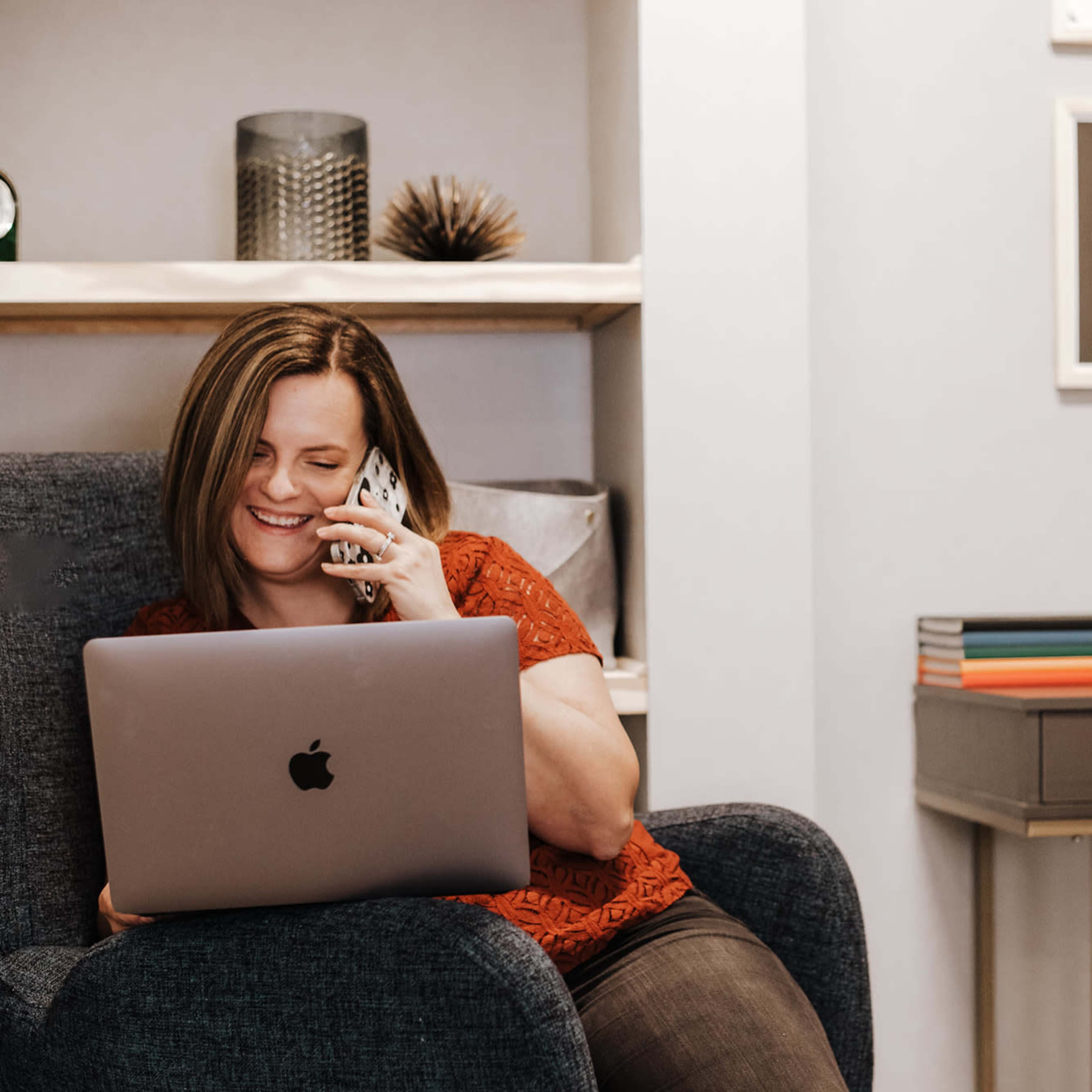 A woman is sitting in a chair, using a laptop while talking on a smartphone.