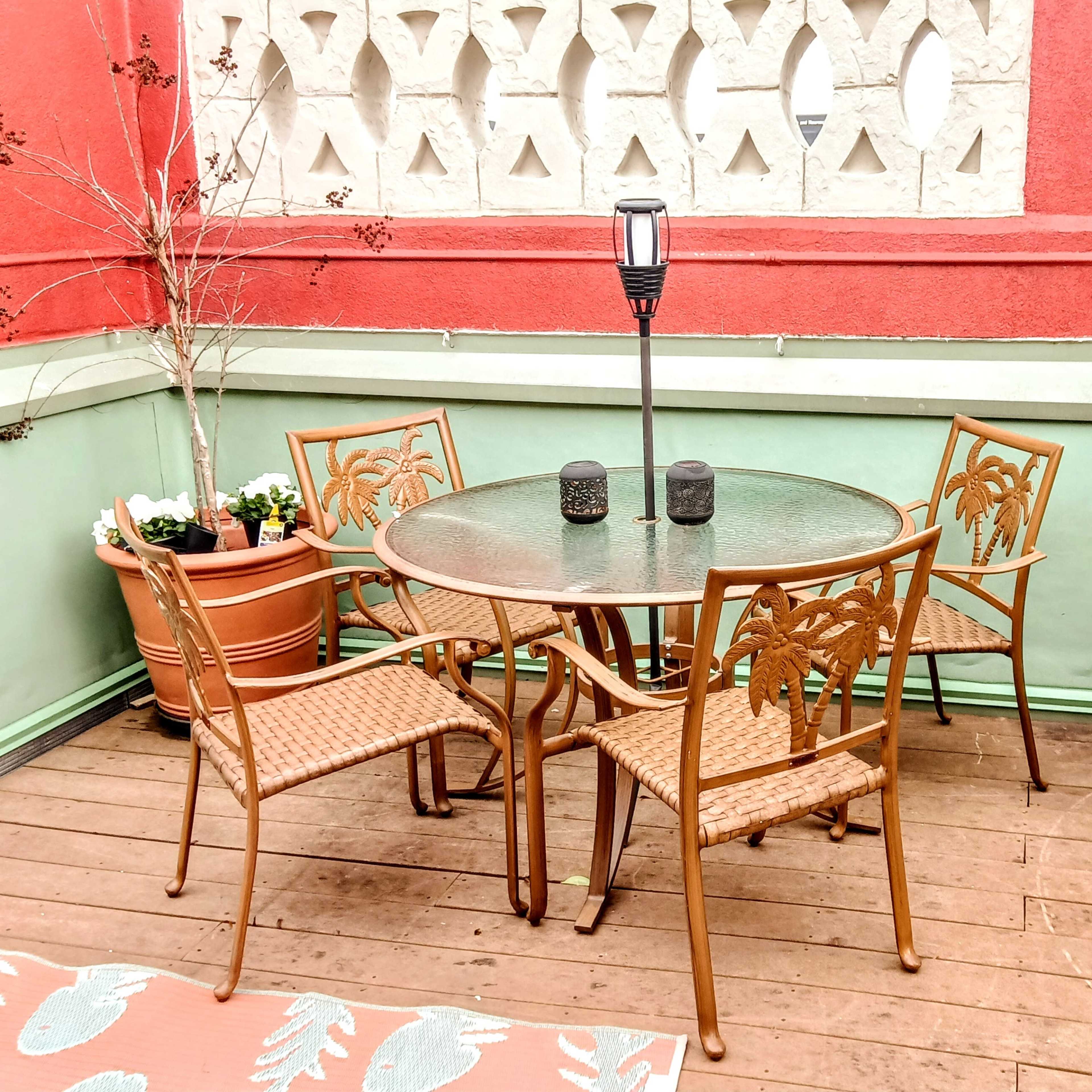 A patio features a circular glass table surrounded by four chairs, with potted plants and a lantern nearby.