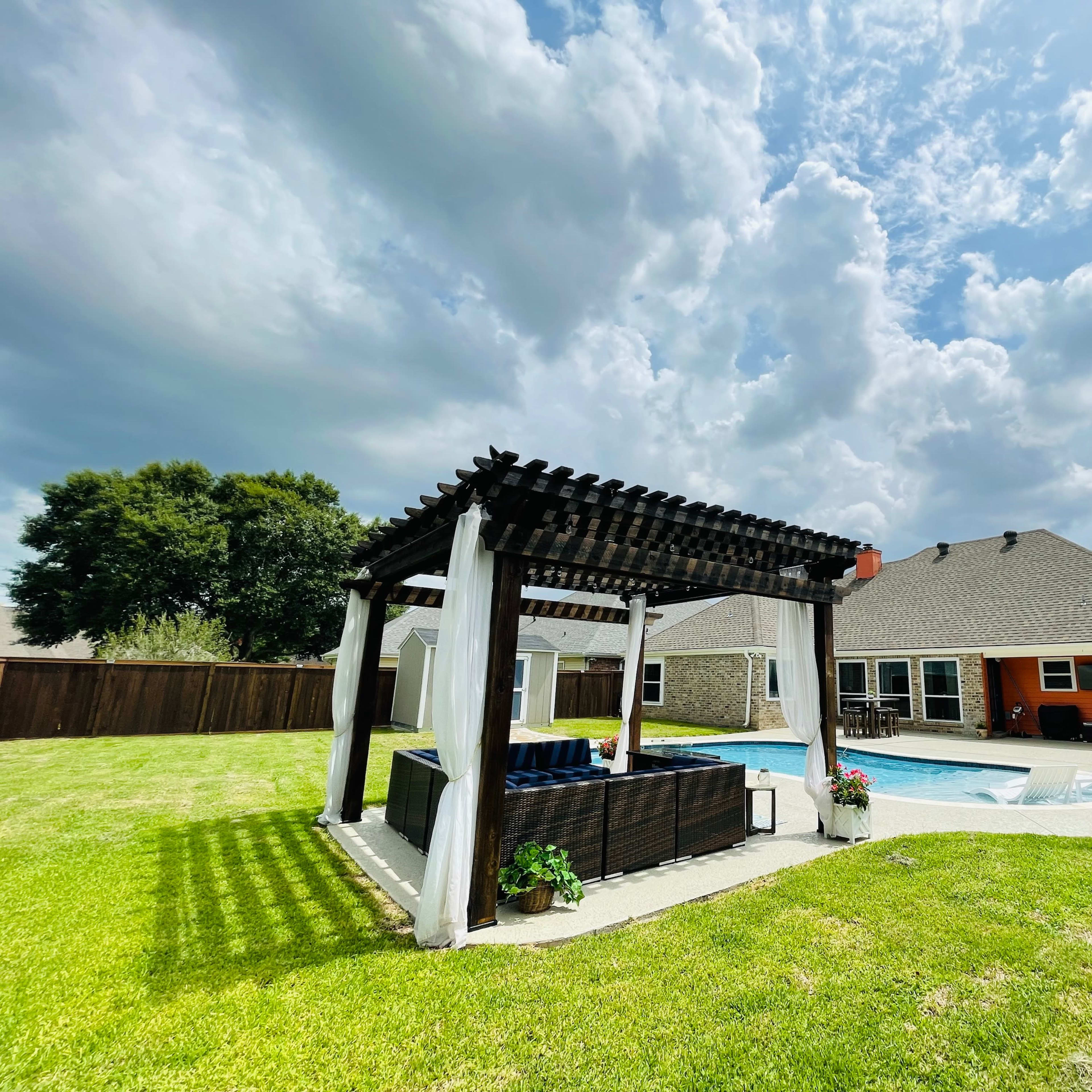 A backyard featuring a pool surrounded by grass, a covered seating area, and a house with a mix of exterior colors.