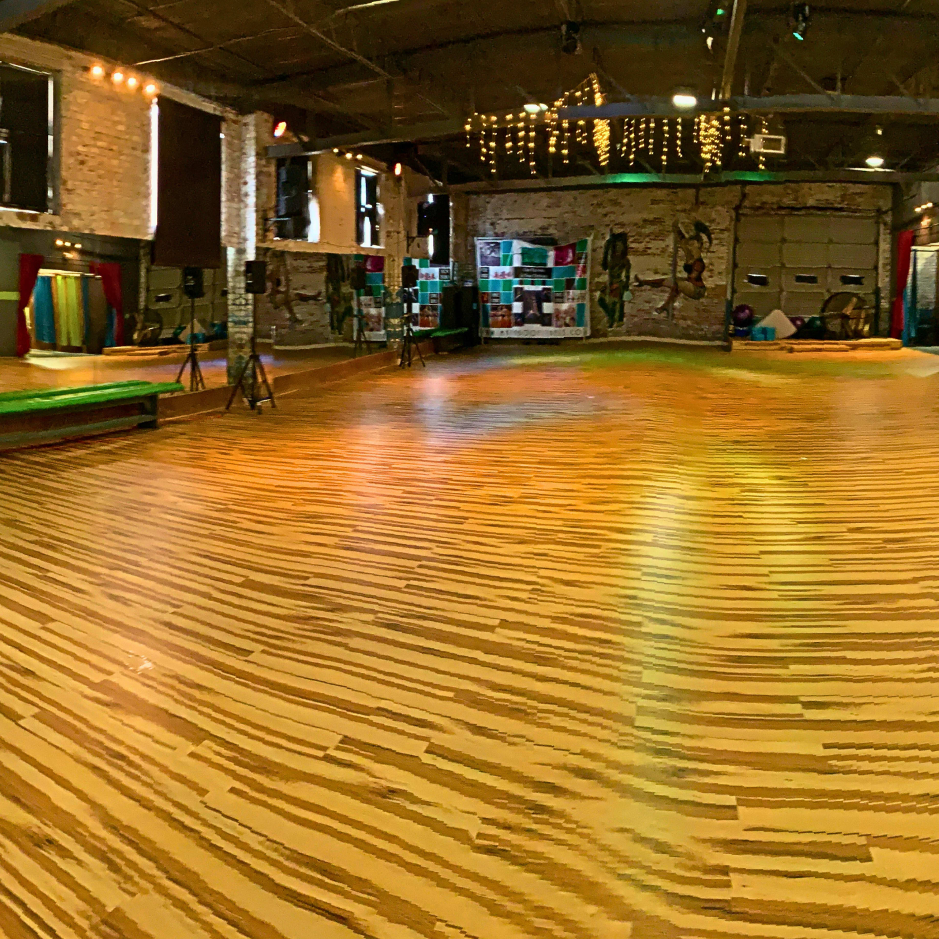 A spacious wooden-floored dance studio with exposed brick walls and various props along the sides.